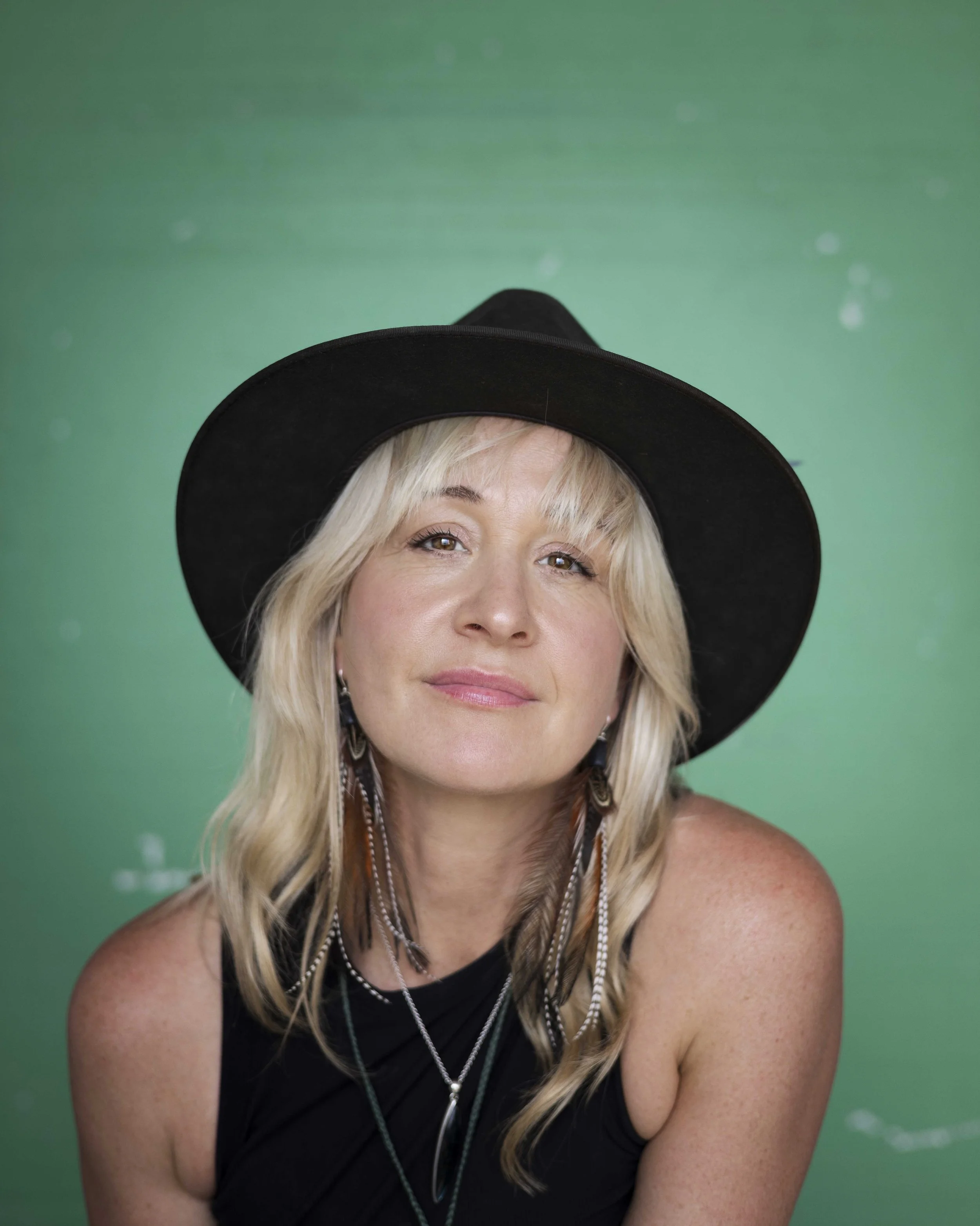 Head and shoulders  staff portrait of Heidi. Green background. Heidi wears a black wide brimmed hat, blonde hair, black singlet and is smiling warmly at the camera.