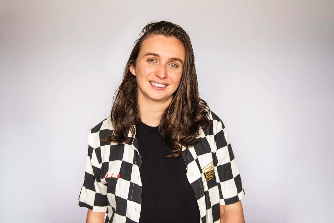 Headshot portrait of Theresa Harrison. Theresa is standing in front of a white background. Brightly lit. Brown wavy shoulder length hair, smiling with green eyes. They wear a black and white checkered shirt.