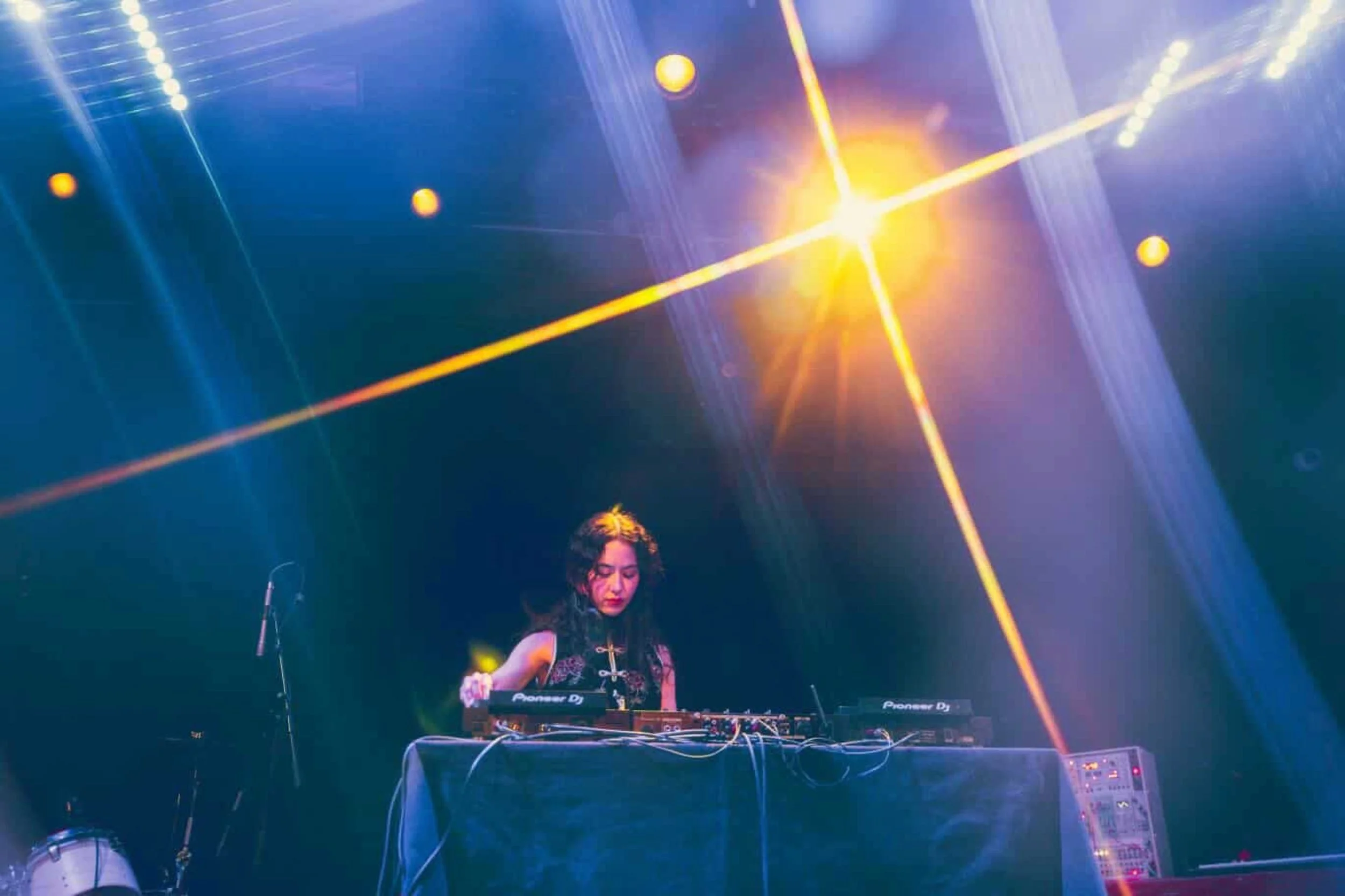DJ Shallowhalo performing live in Dimes Square, on stage with Pioneer DJ decks, dramatic blue stage lighting and golden light flare
