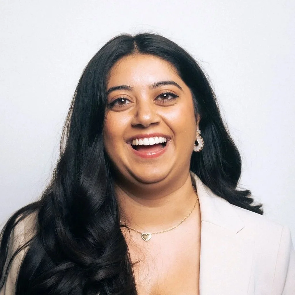 A woman with long black hair, smiling with teeth showing, wearing earrings and a necklace with a heart pendant, dressed in a light-colored blazer against a plain background.