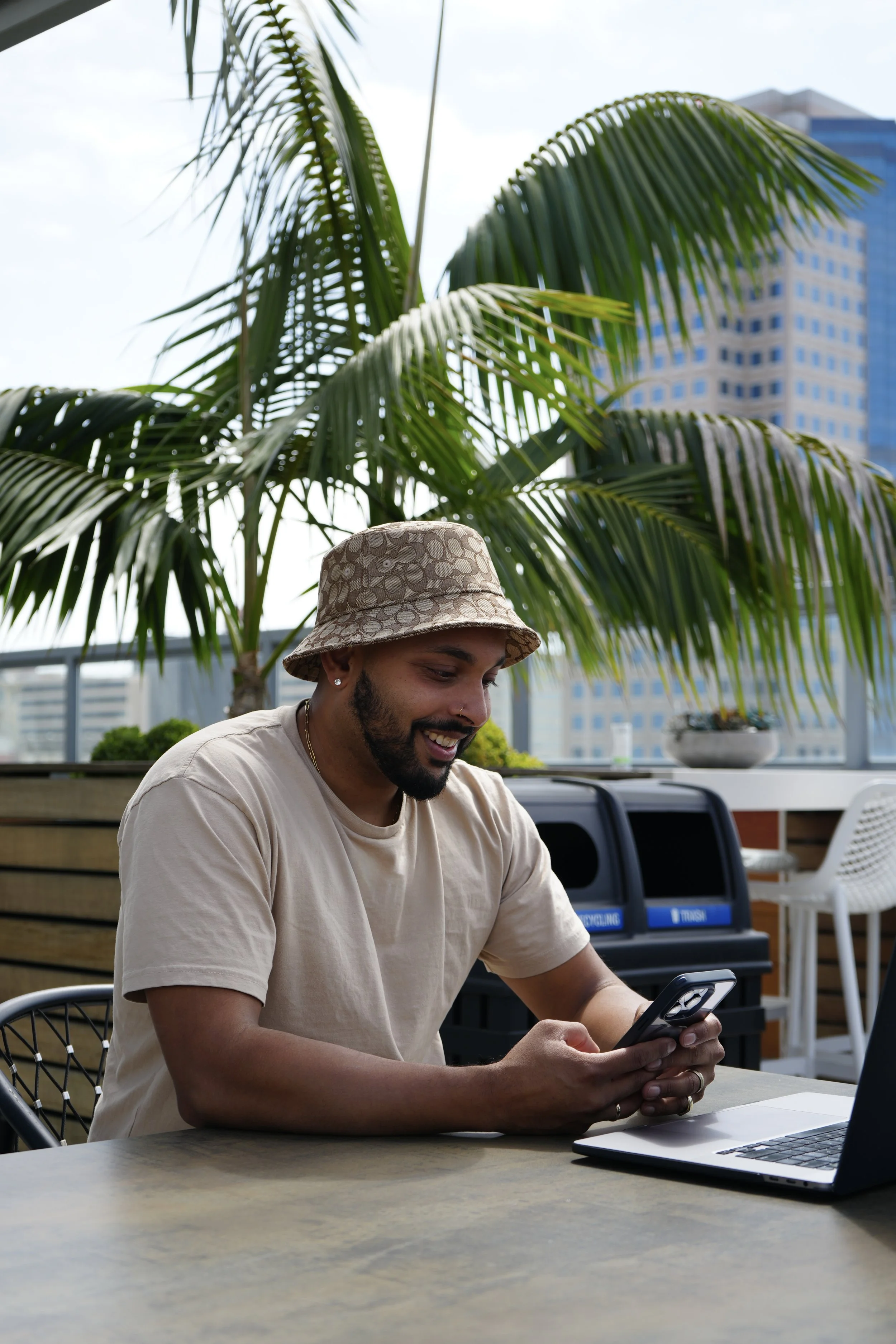 A man smiling while looking at his phone, sitting at a table on a rooftop terrace with a laptop nearby, greenery, and tall buildings in the background.