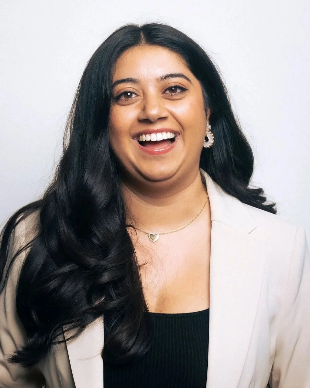 A woman with long black hair, wearing earrings and a delicate necklace, smiling widely and showing her teeth, dressed in a light-colored blazer over a black top.