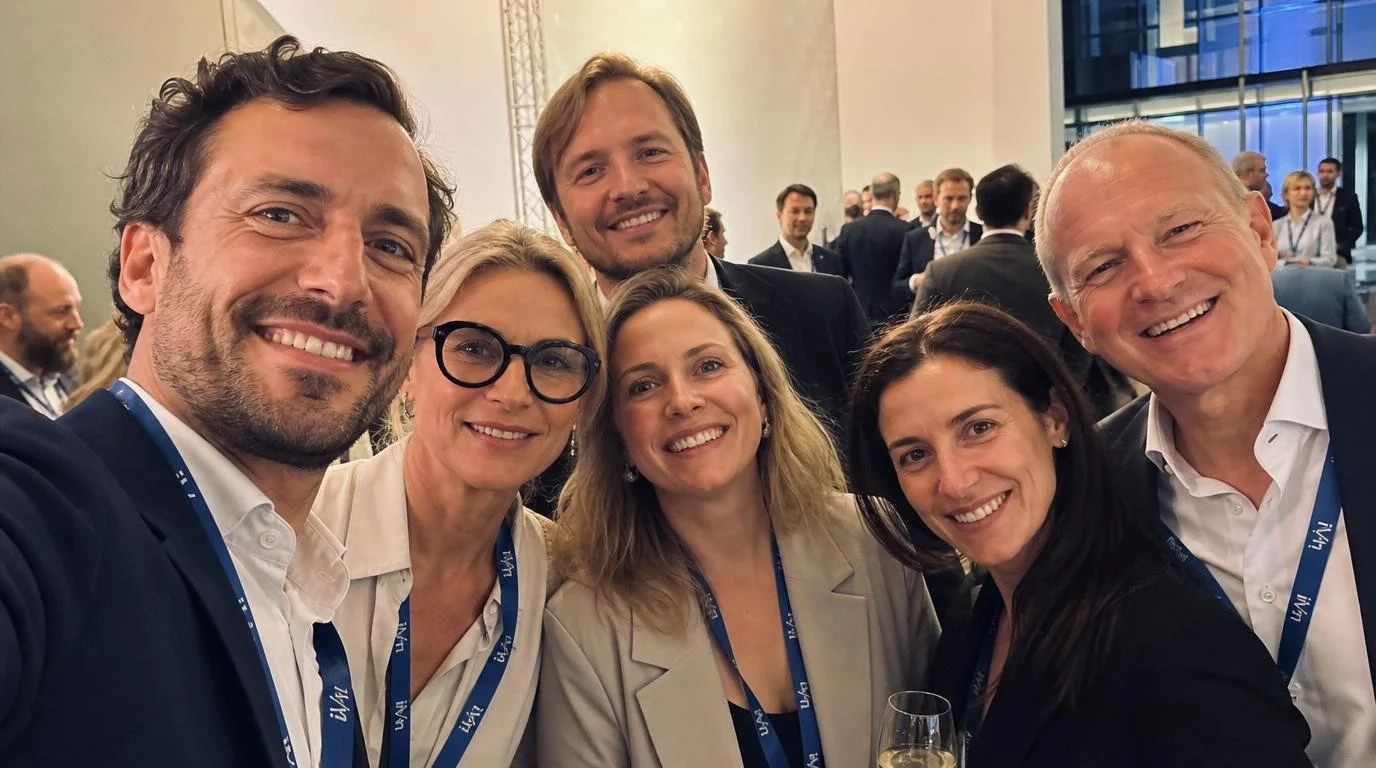 Group of six smiling people taking a selfie at a professional event or conference, with name badges and a busy background.