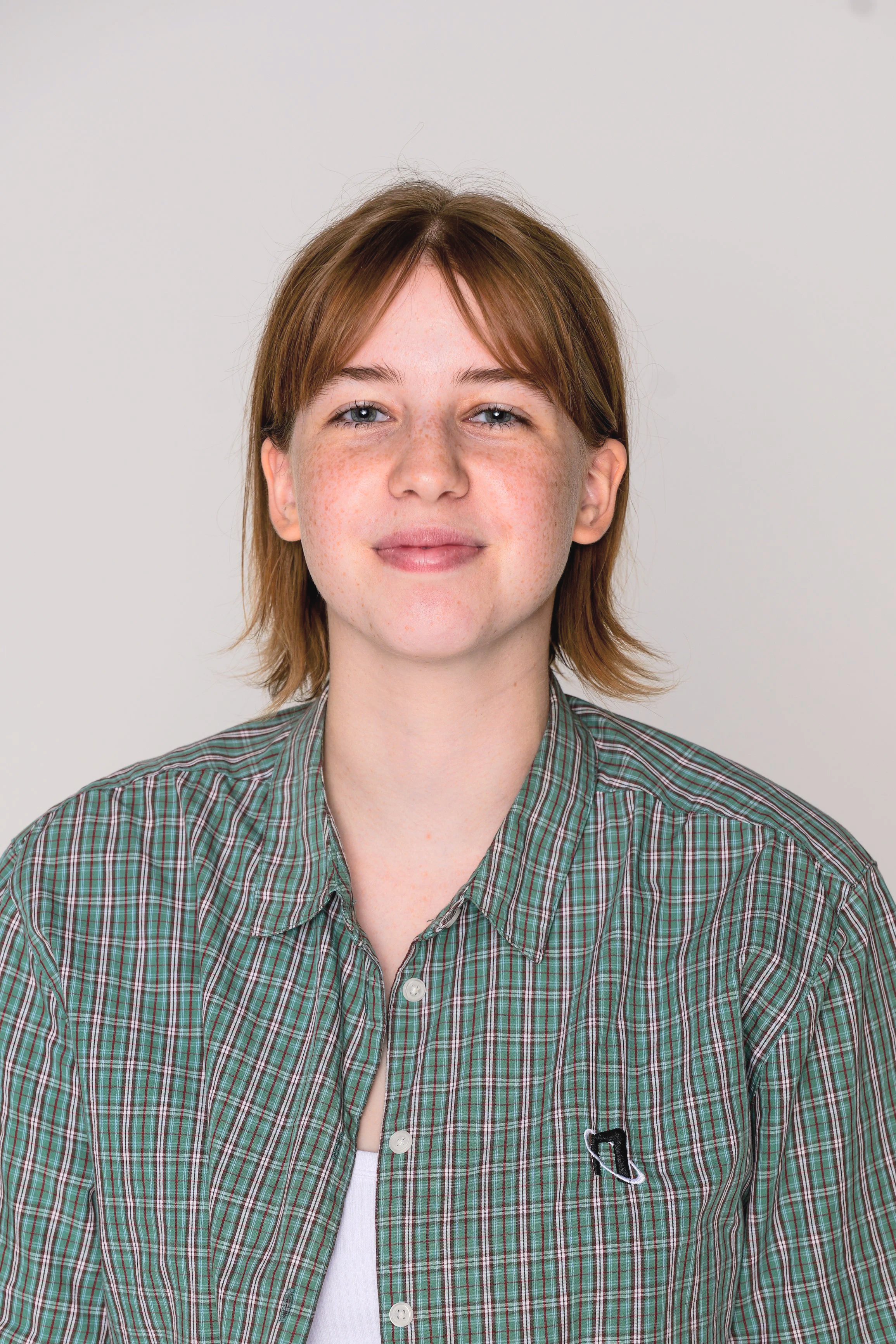 A young person with short red hair, light skin, and freckles, wearing a green plaid shirt and smiling slightly, standing against a plain light background.