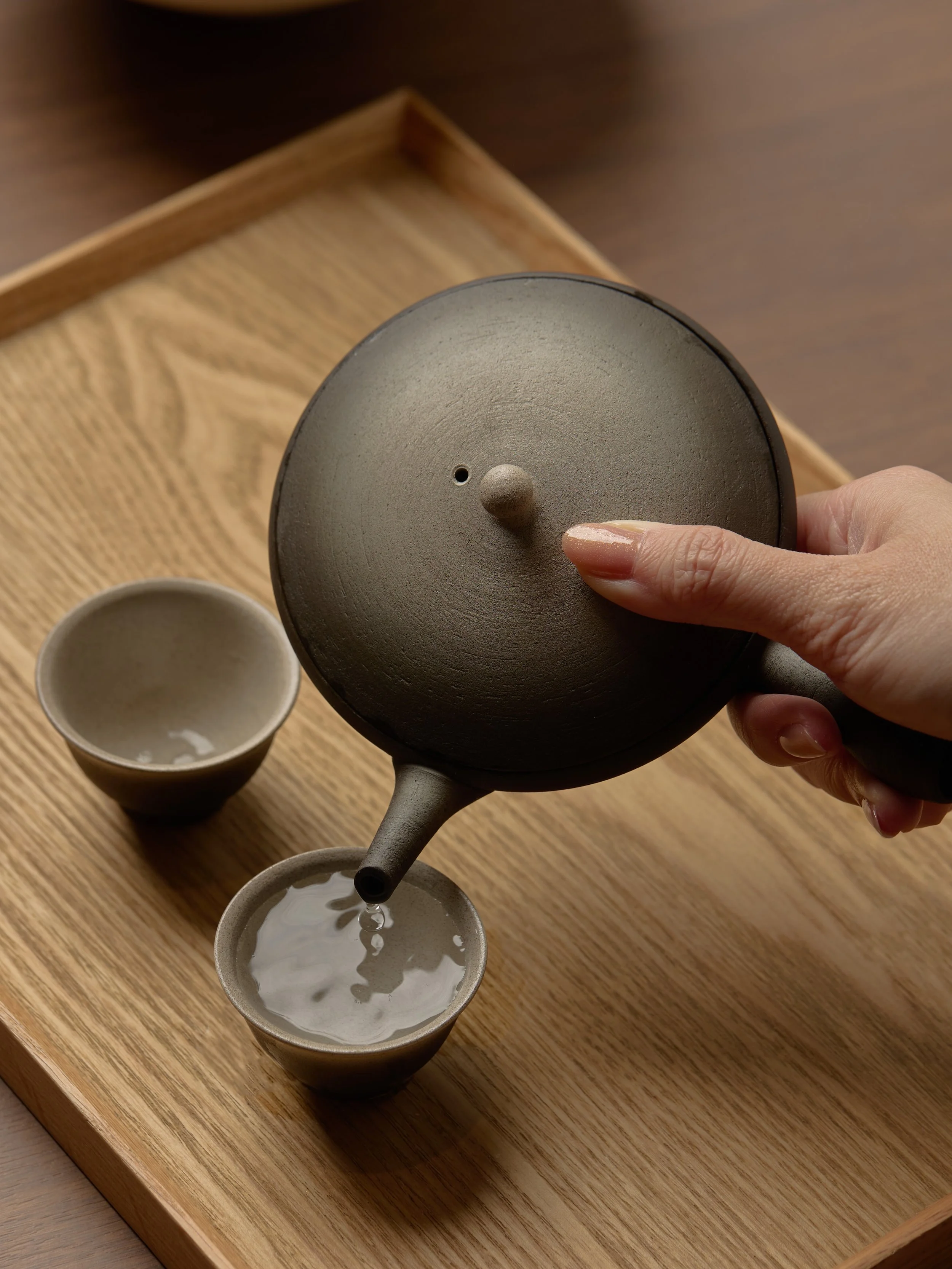 Person pouring tea from a dark-colored teapot into two small matching cups on a wooden tray.
