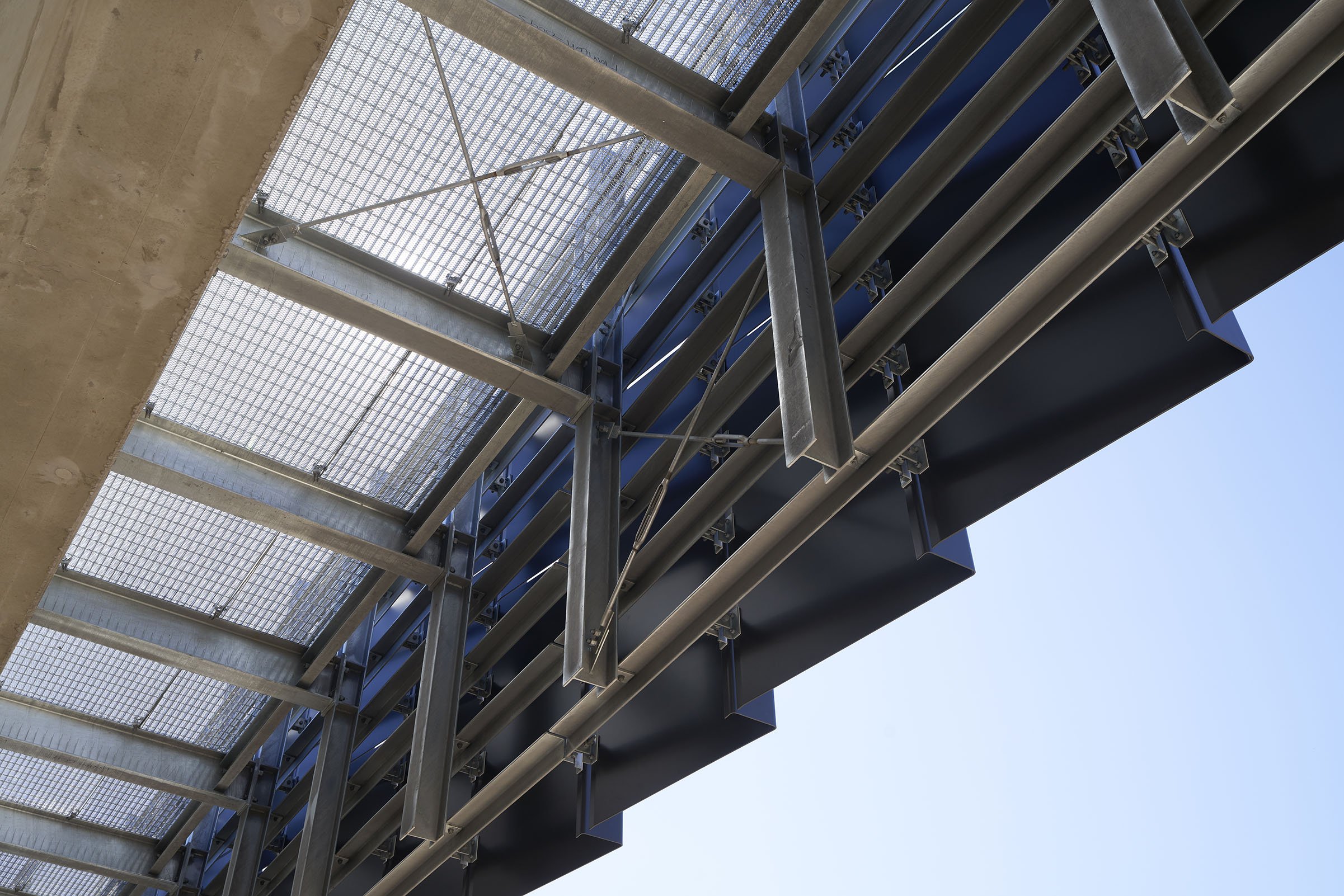 View of a construction or industrial structure with metal beams, guardrails, and a metal mesh platform against a clear blue sky.