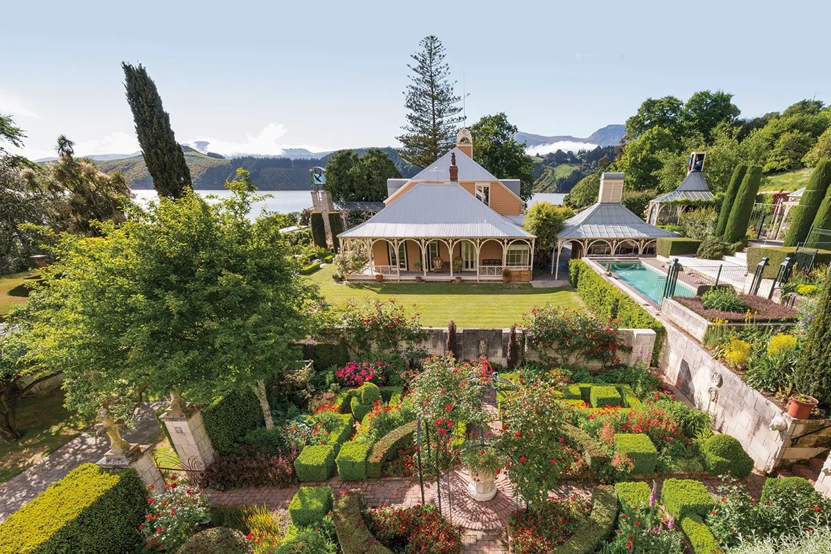 A lush garden with colorful flowers, green hedges, and trees, with a house and swimming pool in the background near a body of water and mountains.