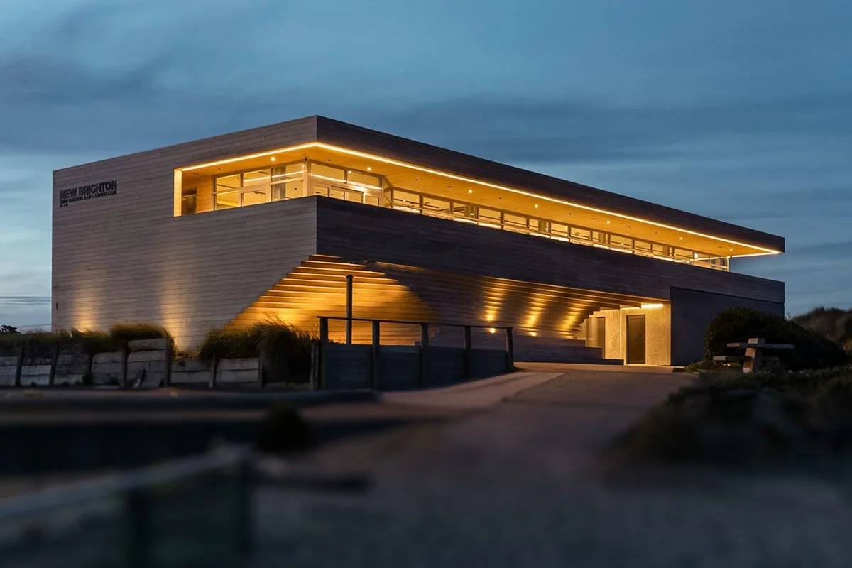 Modern multi-story building with wooden exterior, illuminated at night, with large windows and an outdoor staircase leading to a balcony.
