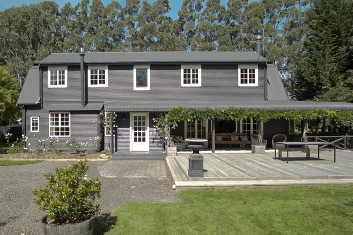 A two-story house painted in dark gray with white window frames and a gray roof, featuring a front porch with benches and a pergola covered with green vines, a paved walkway, and a grassy yard surrounded by trees.