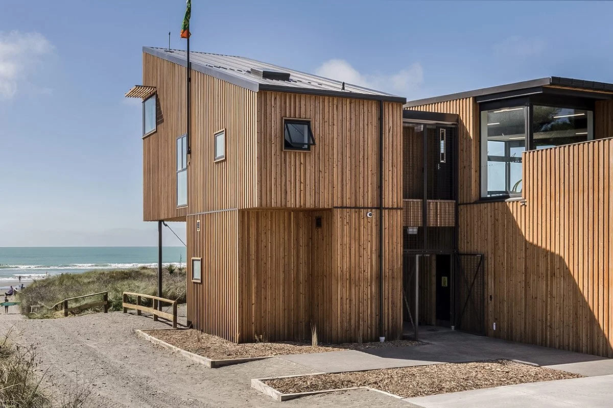 Modern wooden beachfront house with multiple levels and large windows, located near the beach with sandy pathways and dunes.