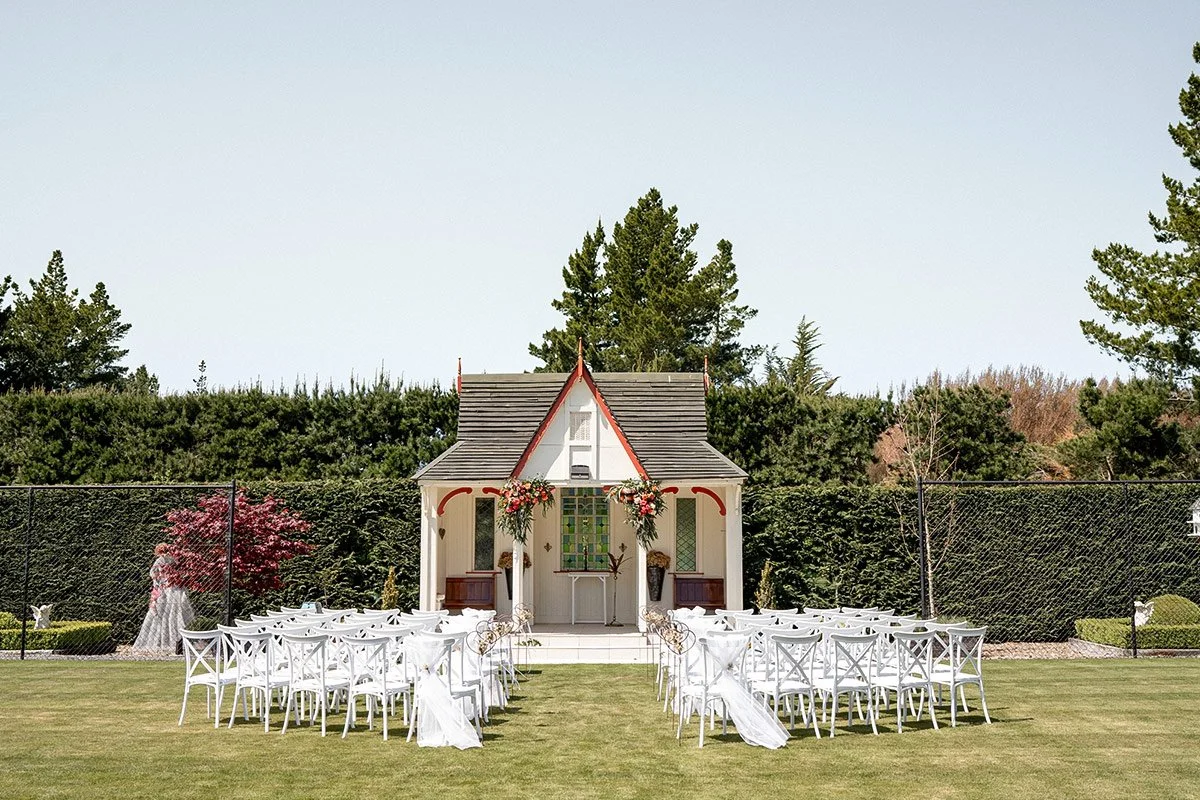 Outdoor wedding setup with white chairs and small chapel surrounded by green bushes and trees.