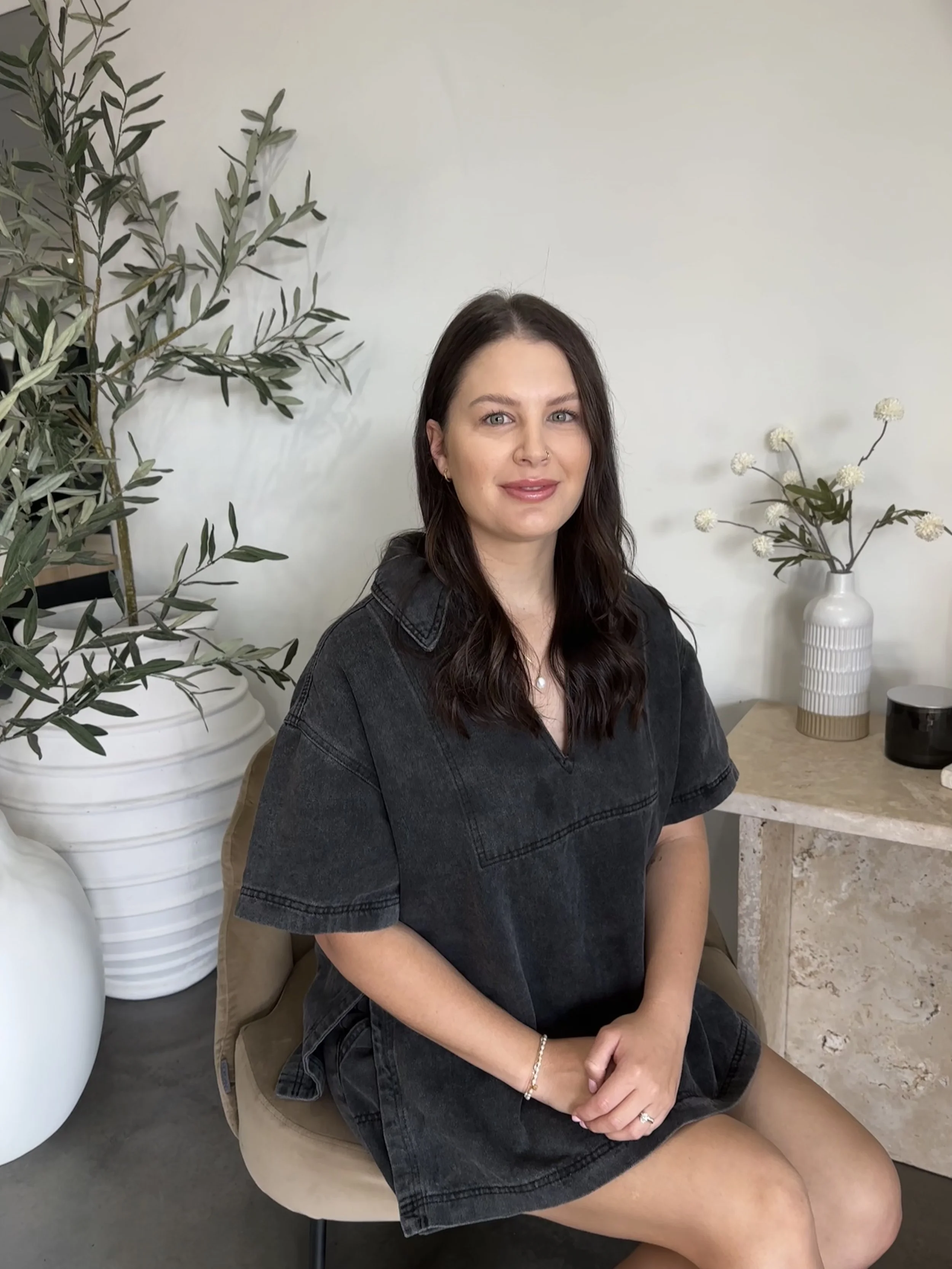 A woman with long dark hair wearing a black dress sitting on a beige chair in a modern room with large white vases, green plants, a marble side table, and floral arrangements.