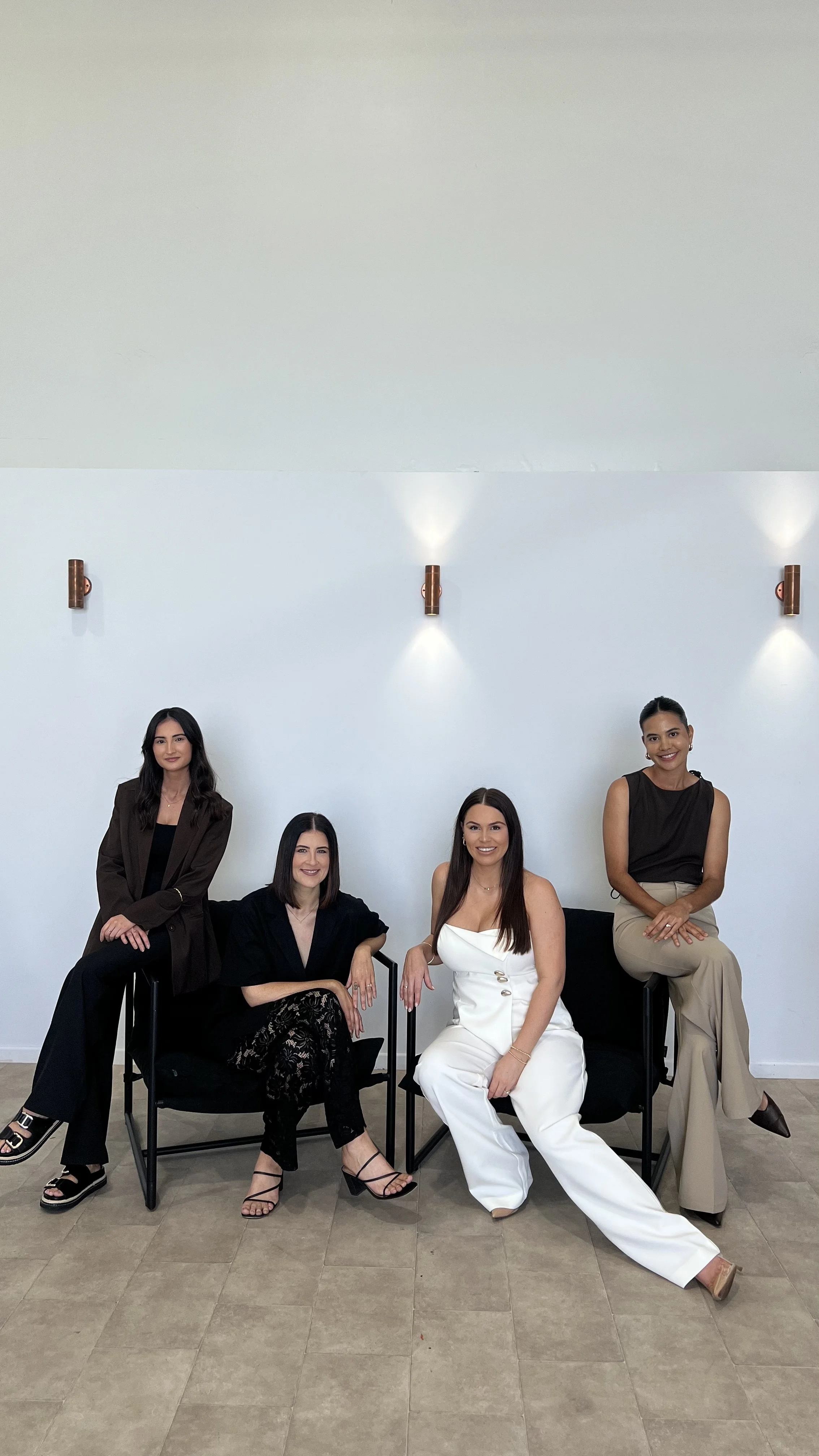 Four women with dark hair and light to medium skin tones posing with smiles in stylish business casual attire in front of a plain white wall with modern wall-mounted lights.