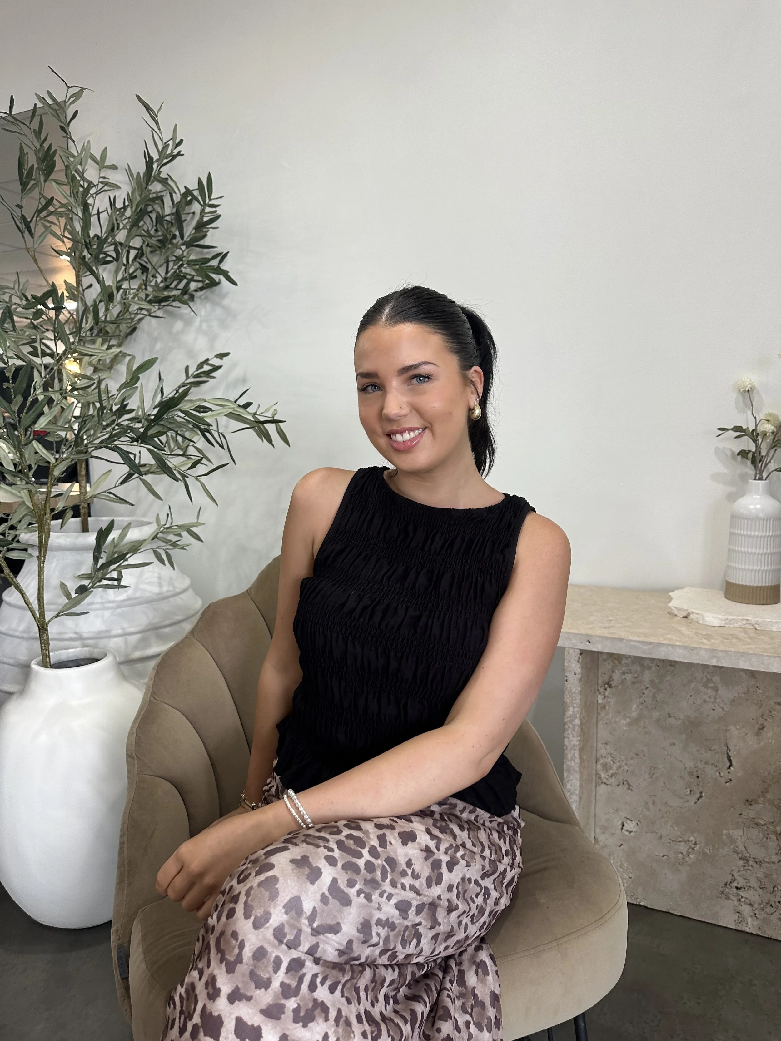 A young woman with shoulder-length blonde hair, wearing a beige sleeveless jumpsuit, sits casually on a chair with black cushions in a minimalistic room with white walls and tile flooring. She is smiling and looking at the camera.