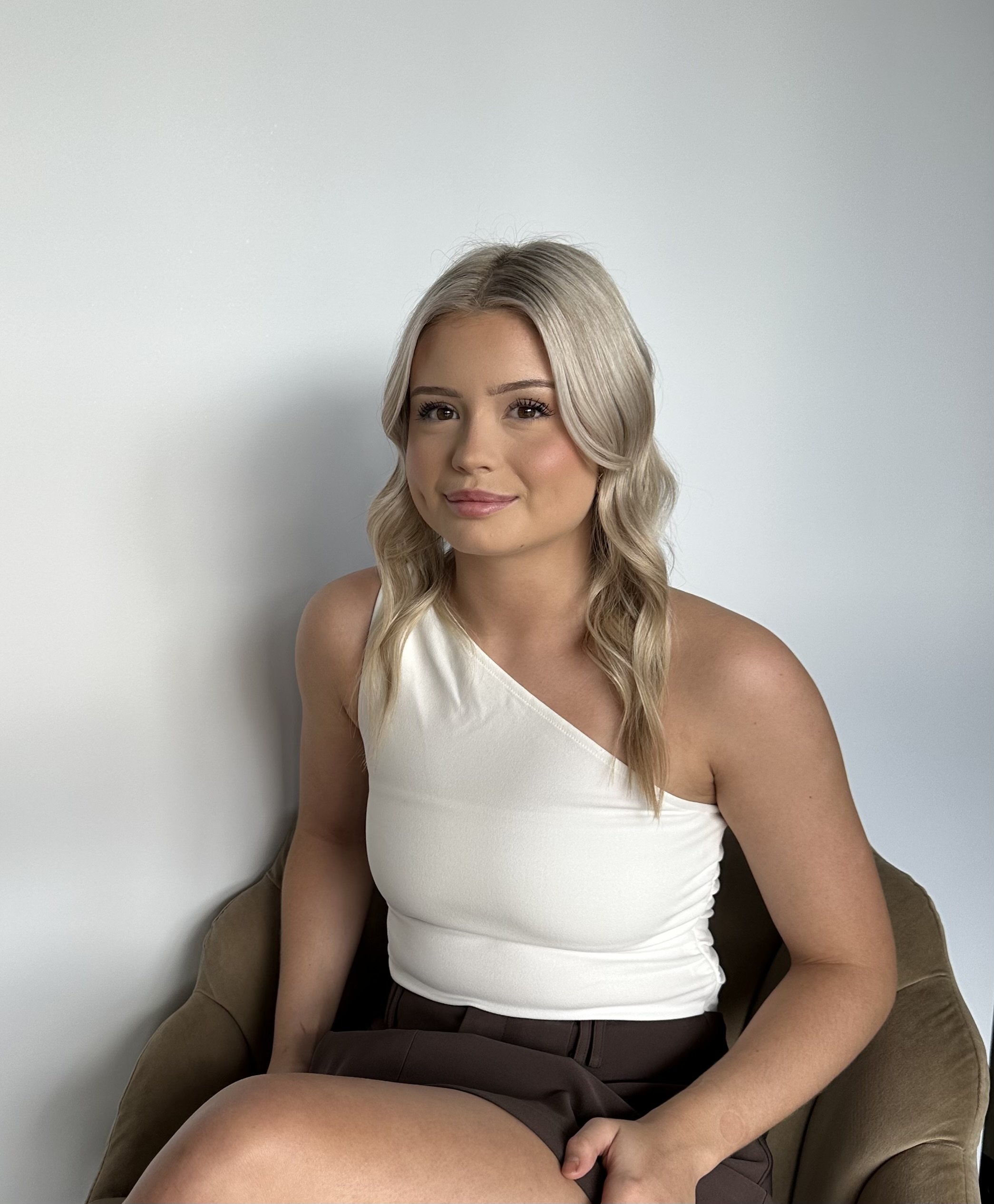 A smiling young woman with blonde hair sitting on a black chair. She is wearing a black sleeveless top and cream-colored wide-leg pants, with tattoos on her arms. She is in a minimalistic room with white walls and tiled flooring.