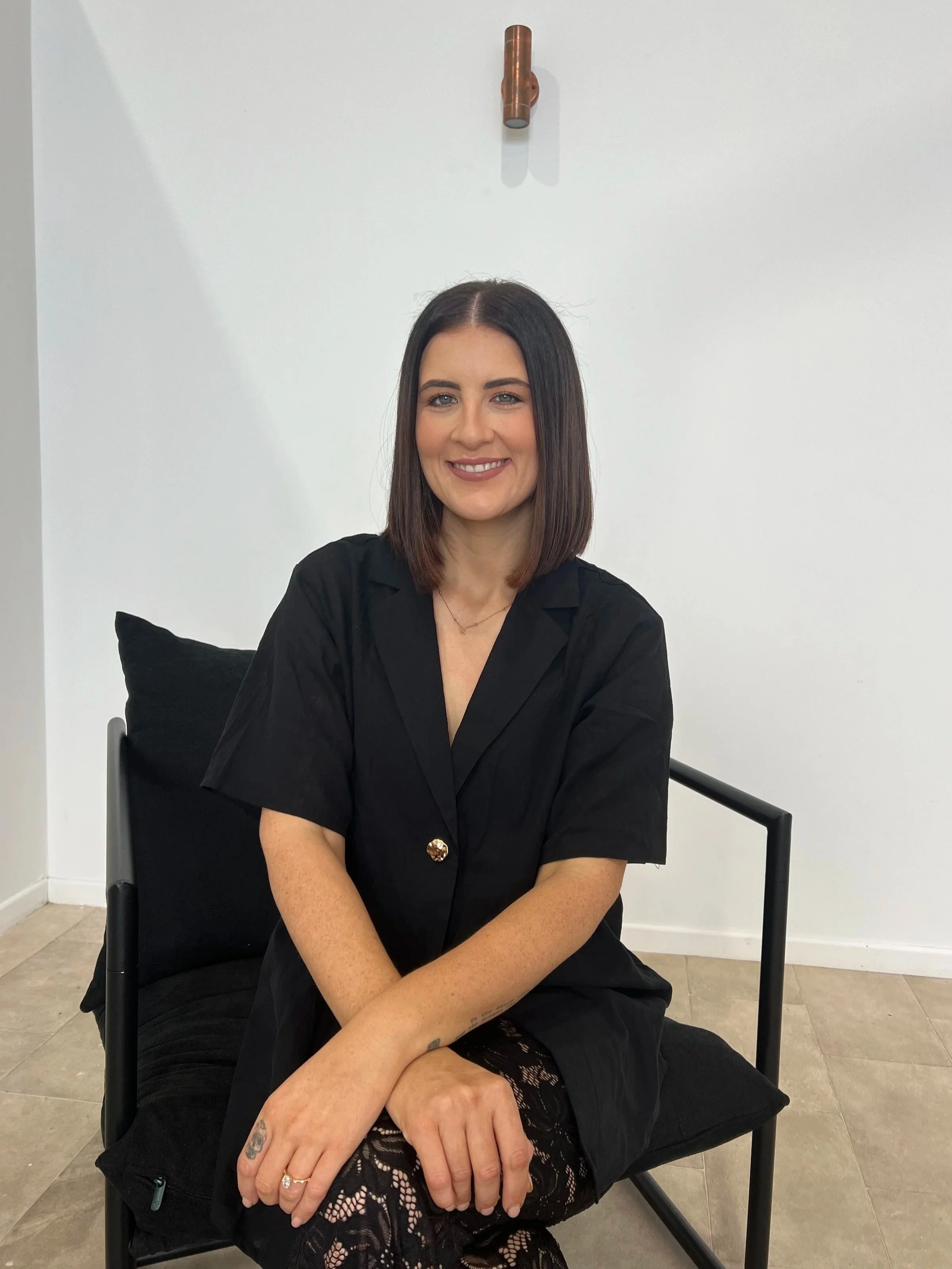 A woman with shoulder-length dark hair and light skin is sitting on a black chair with a pillow, smiling at the camera, wearing a black dress with lace stockings, in a minimalist room with a white wall and wooden floor.