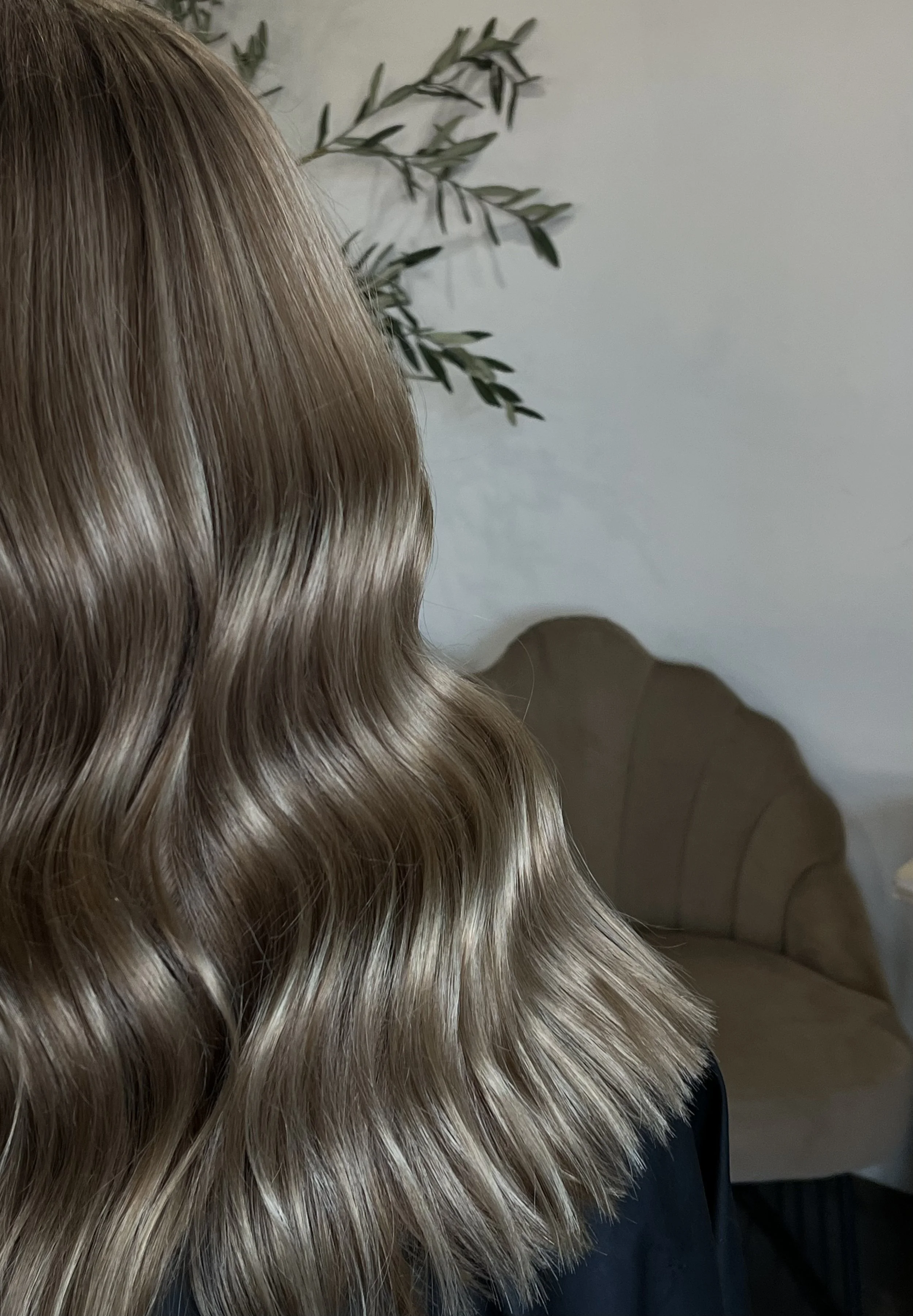 A close-up of smooth, wavy, blonde hair flowing over the shoulder, with a background of a white wall, a green plant, and a beige armchair.
