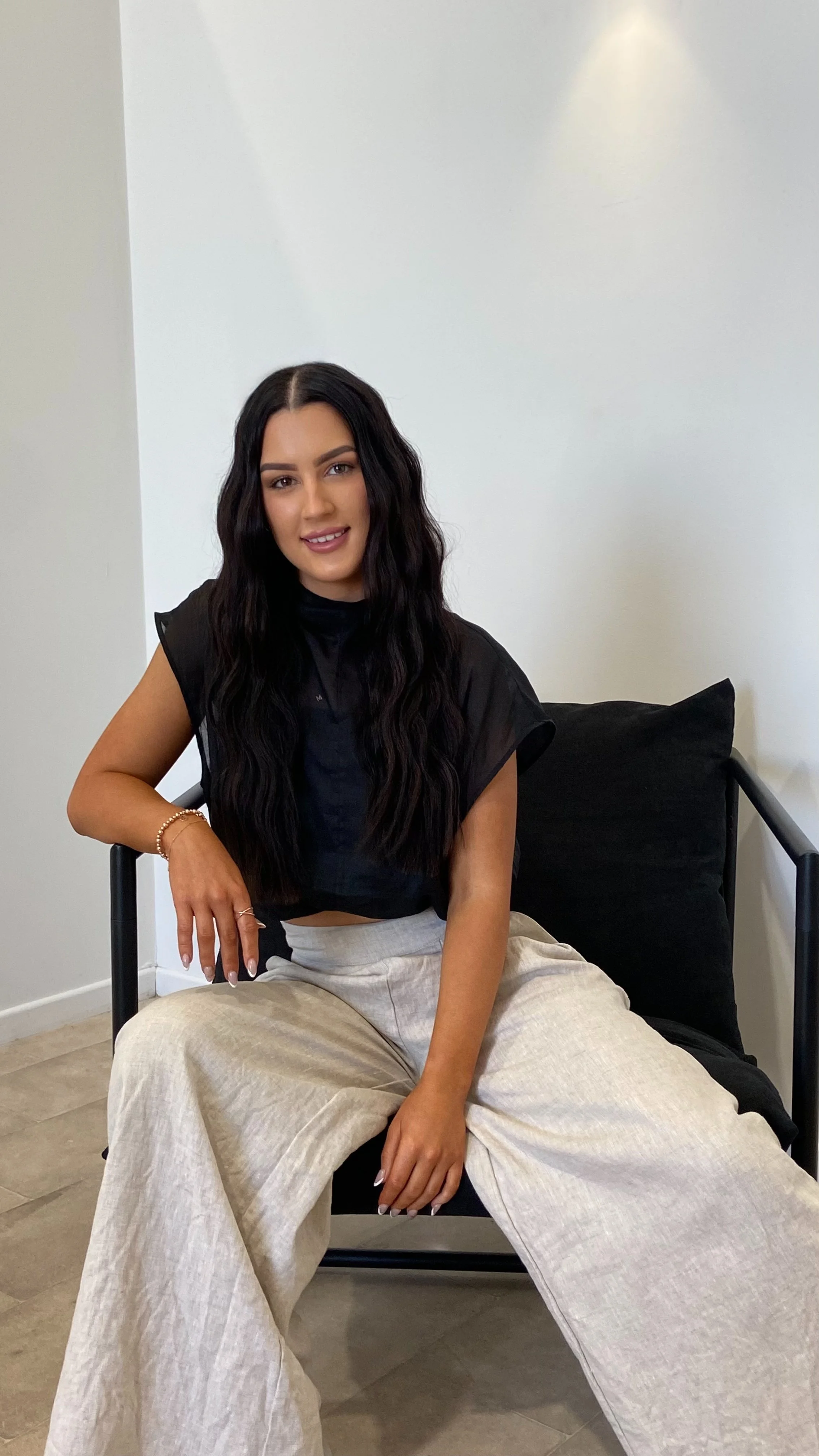 A woman with long black wavy hair sitting on a black chair with black pillow, wearing a black sleeveless top and light beige wide-leg pants, smiling at the camera against a white wall.