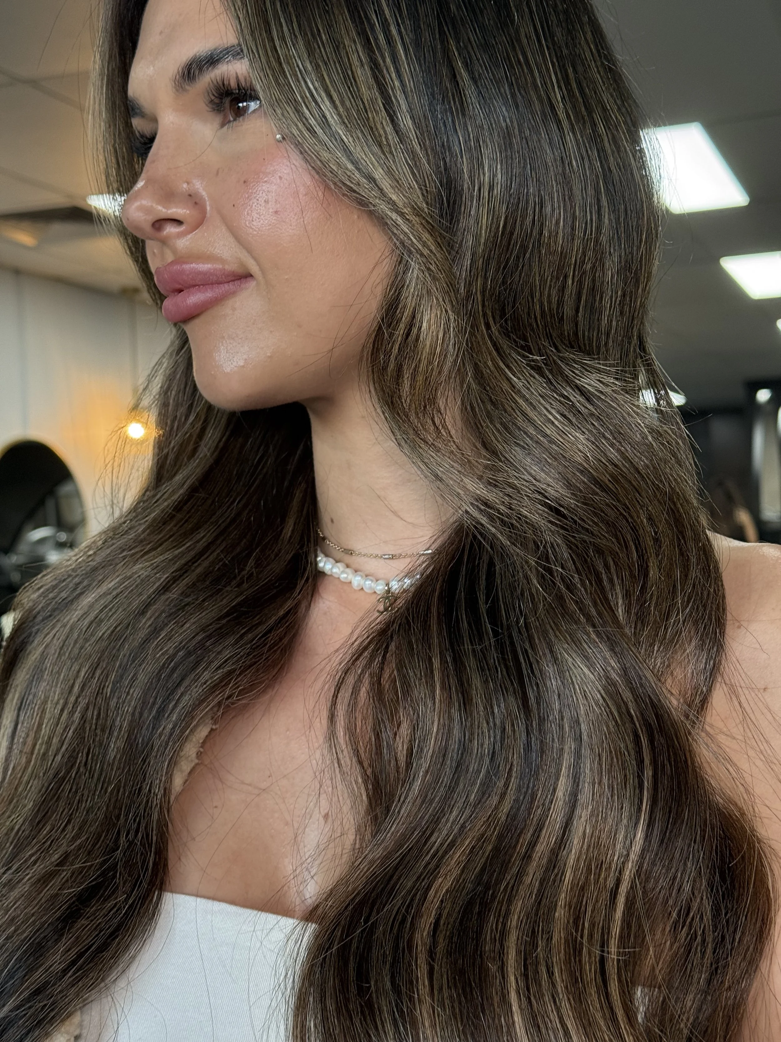 Close-up of a woman with long, wavy brown hair with subtle highlights, wearing a pearl necklace and a white top, in a salon environment.