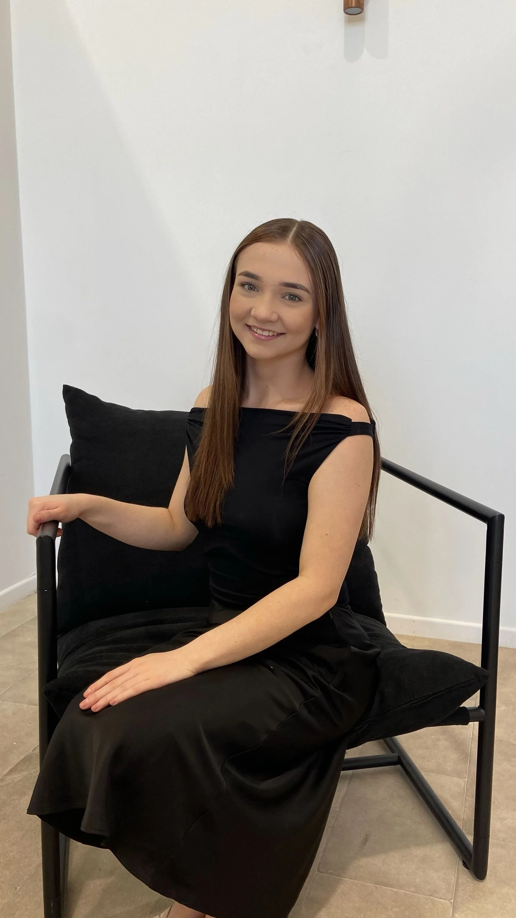 A young woman with long brown hair, smiling, sitting on a black chair with black cushions in a minimalistic room with white walls and tiled flooring, dressed in a black off-the-shoulder top and black skirt.