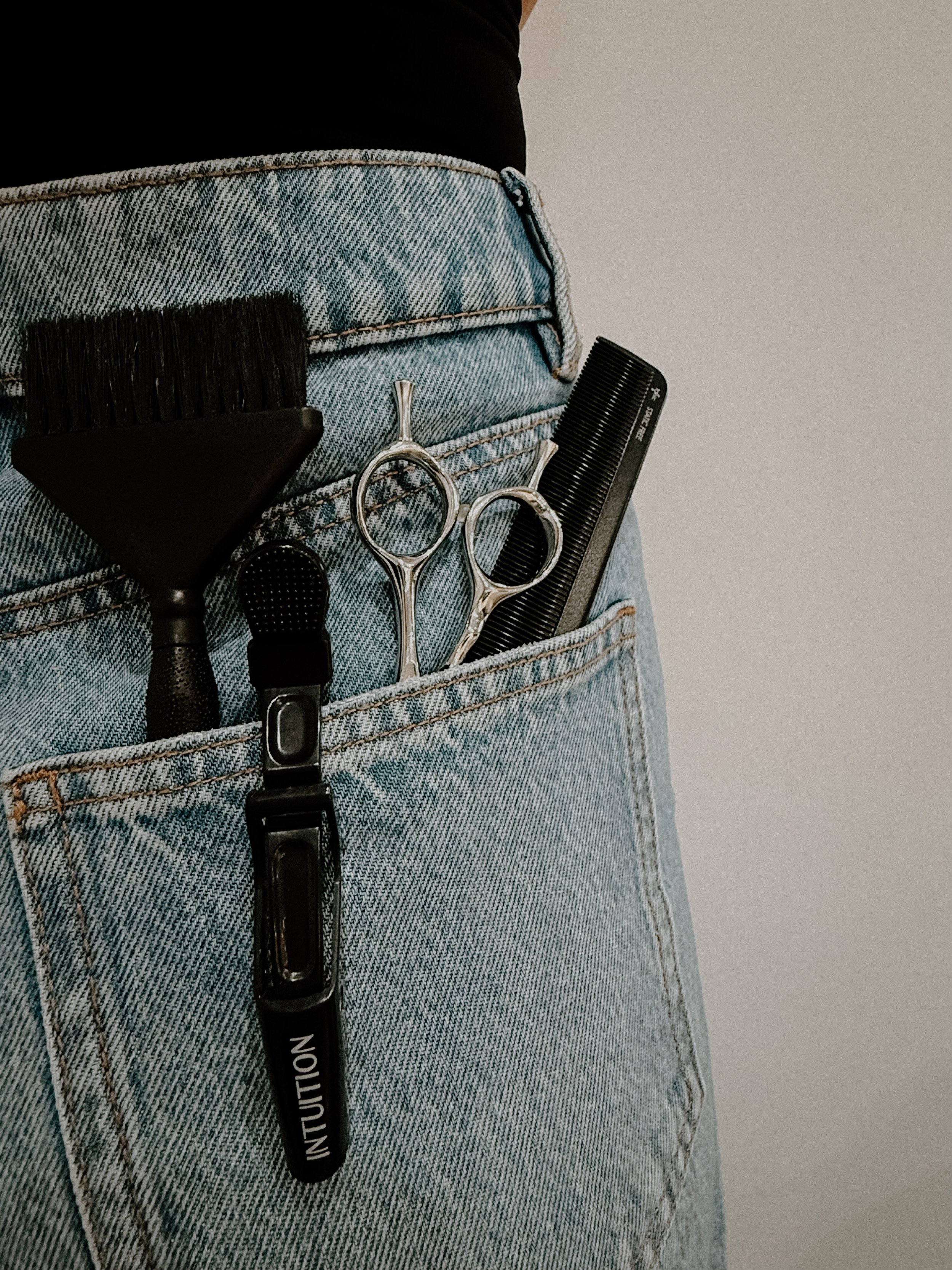 Close-up of a person's back pocket on light blue jeans with hair styling tools inside, including a hairbrush, scissors, a comb, a small brush, and an hair clip with the word "INTUITION".