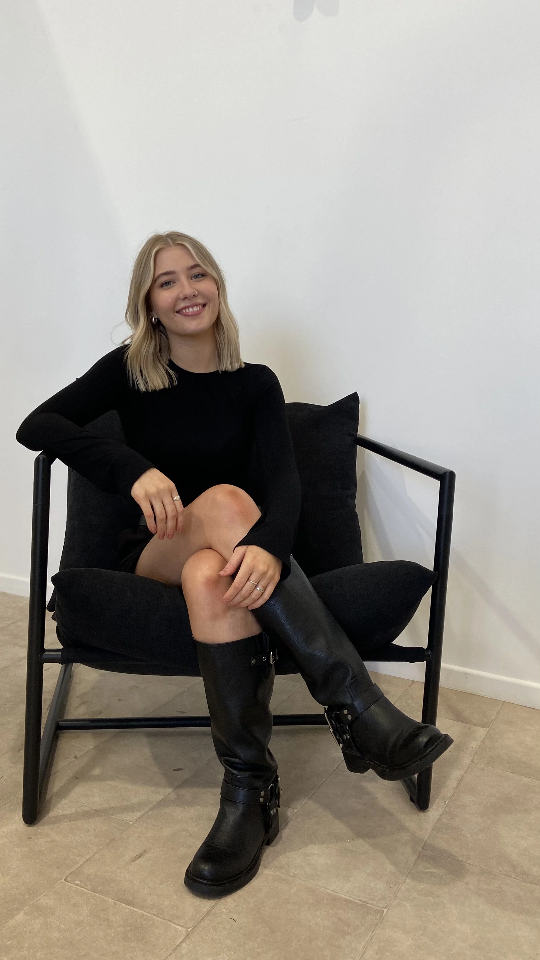 A young woman sitting on a black chair with cushions, smiling and wearing a black dress and black knee-high boots, indoors against a white wall.
