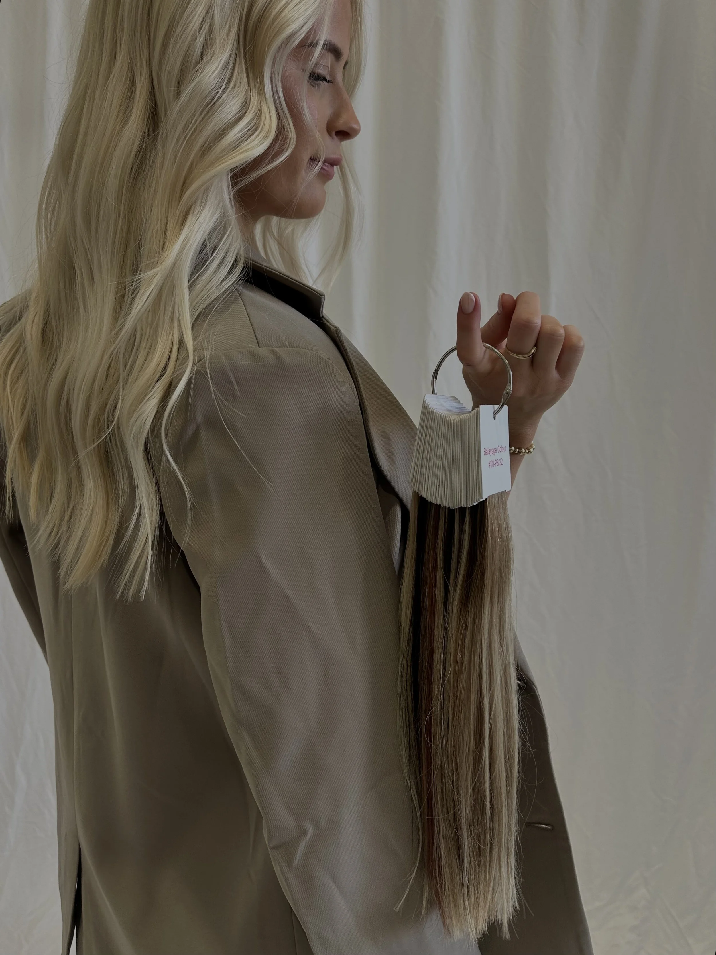 A woman with long blond hair holding a hair extension sample on a ring, standing against a plain background.