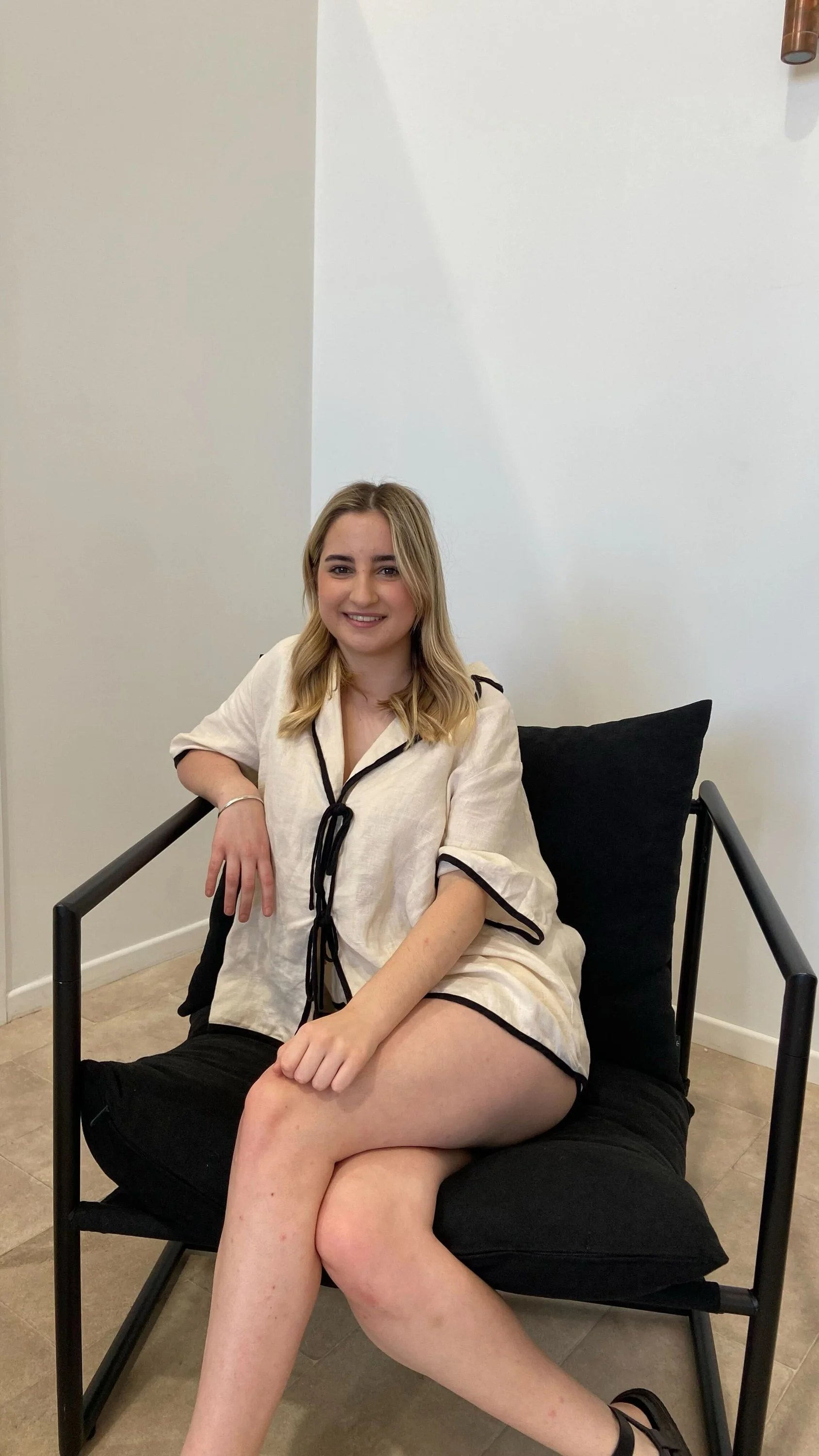 Young woman with blonde hair sitting on a black chair with a black cushion, wearing a cream-colored top with black trim and black shorts, smiling at the camera in a room with white walls and tiled flooring.