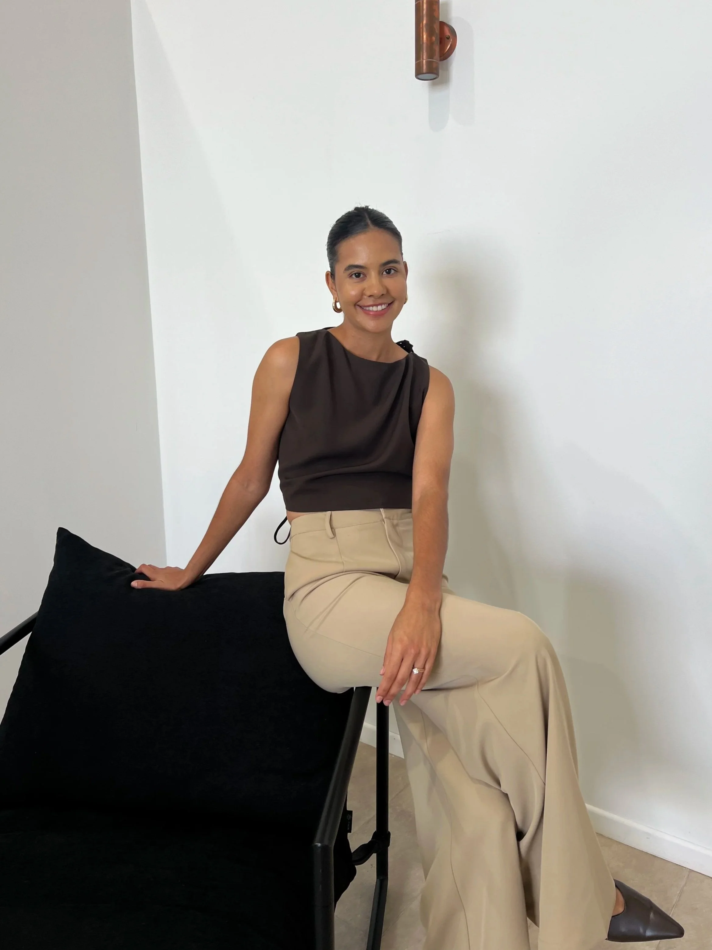 A woman sitting on a black chair with a black pillow, smiling, wearing a sleeveless black top and beige wide-leg pants, with her hair tied back, against a white wall with a copper pipe fixture.