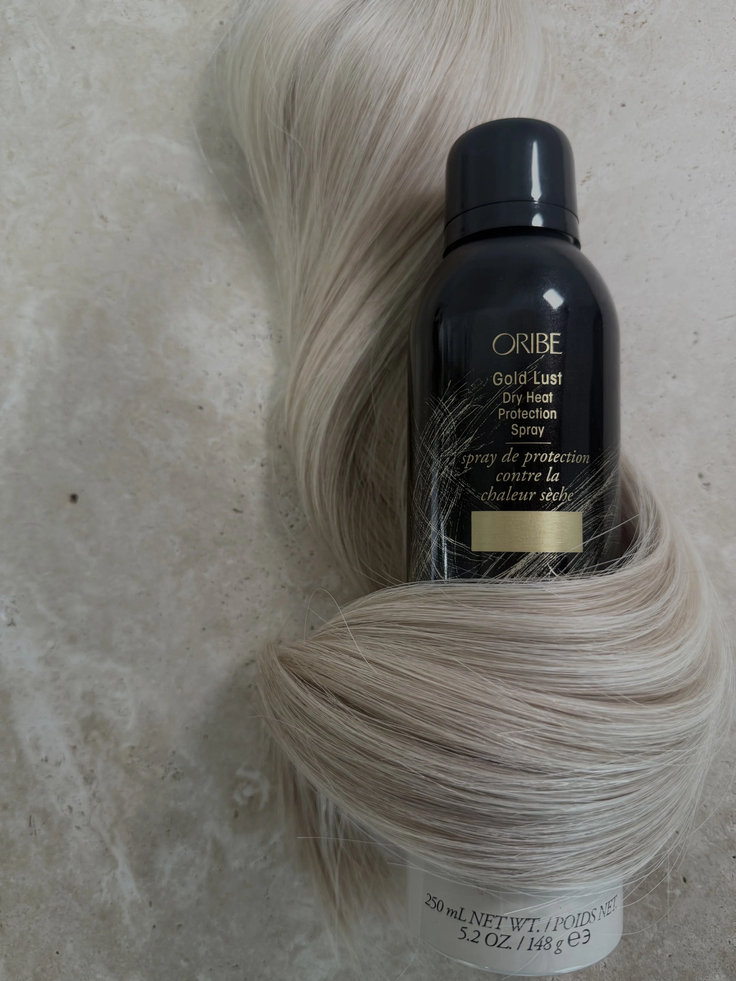 A bottle of Oribe Gold Lust Dry Heat Protection Spray placed on a marble surface with a lock of platinum blonde hair around it.