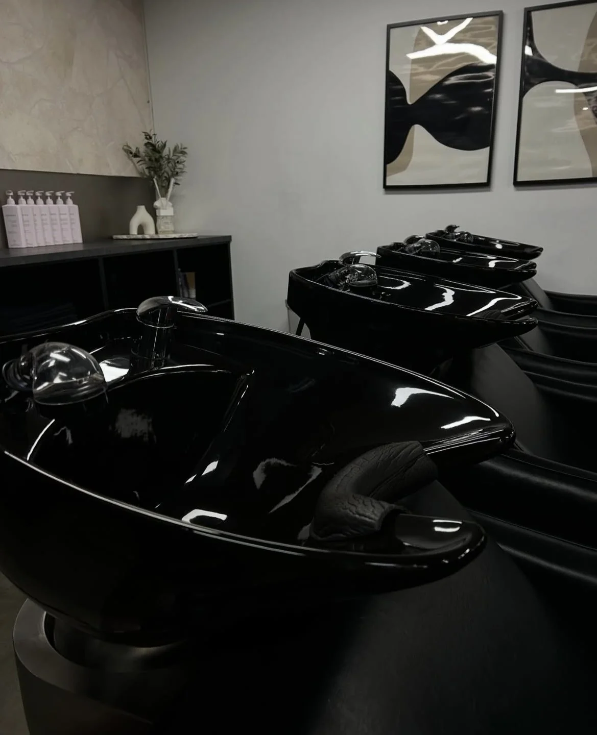 Multiple black hair washing sinks in a salon with wall art and a counter with sanitizer bottles and decorative plants.