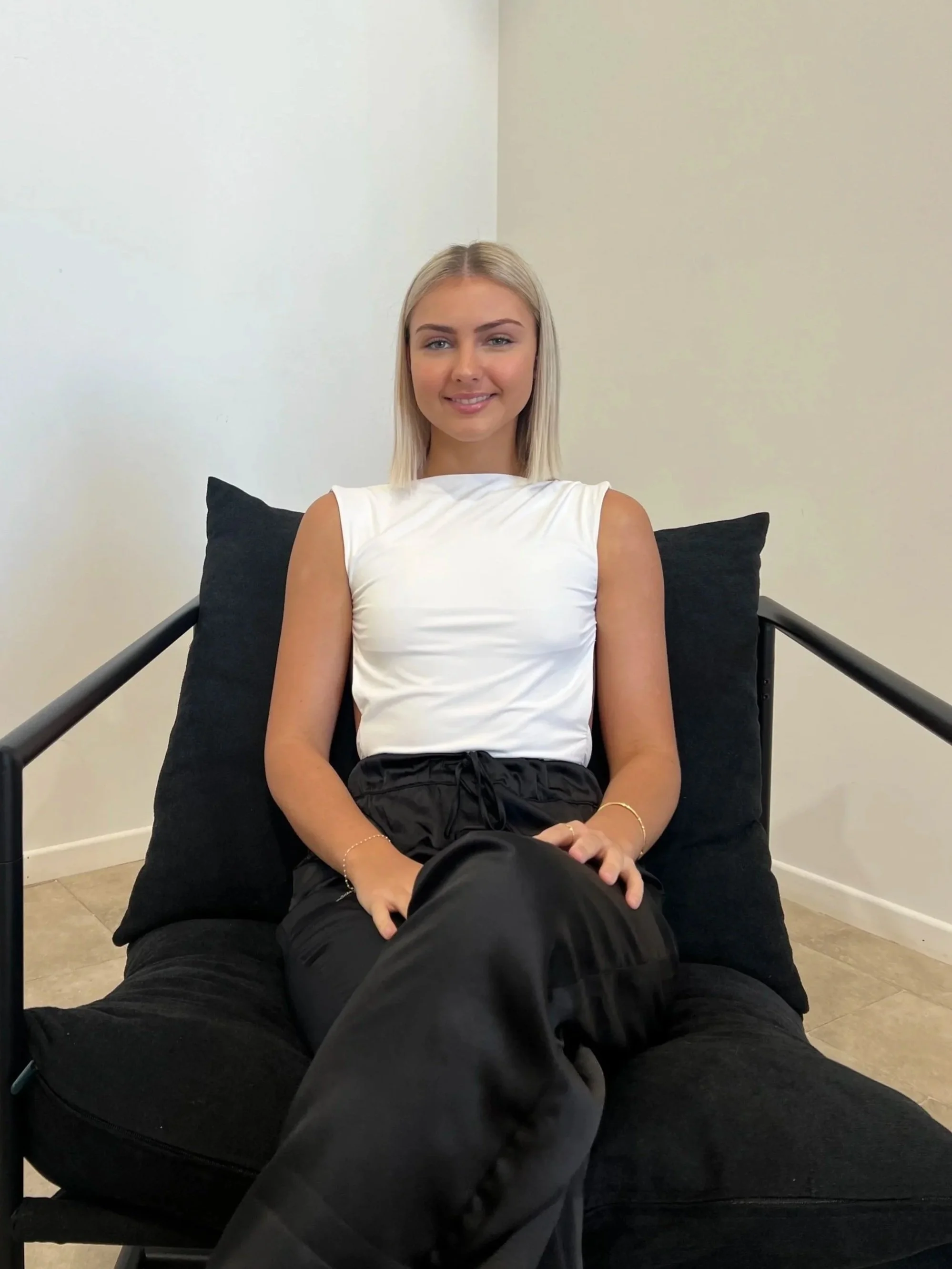 A young woman with blonde hair sitting on a black lounge chair with black pillows, wearing a white sleeveless top and black pants, smiling at the camera.
