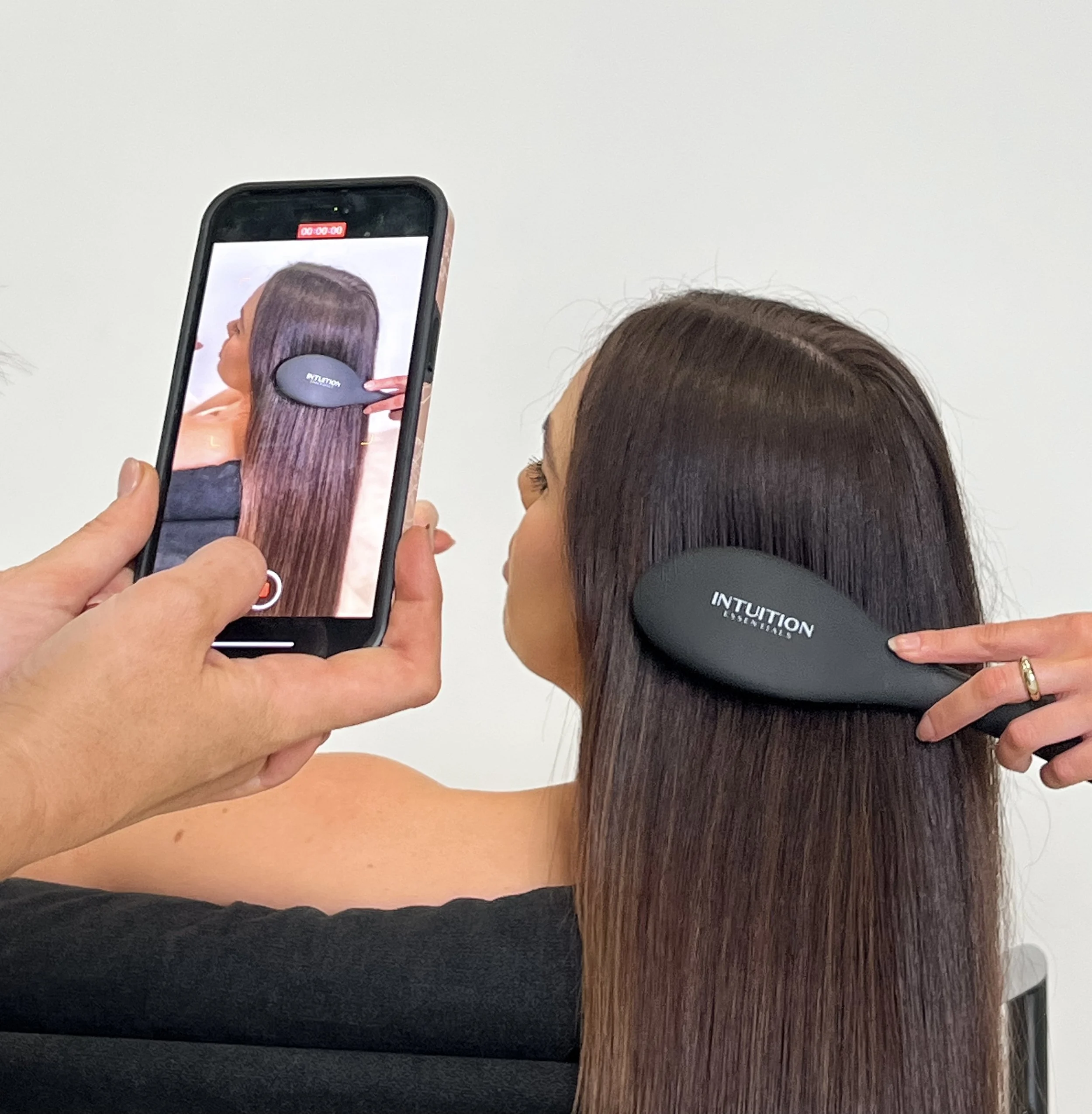 A woman with long, straight brown hair is brushing her hair with a black 'Intuition' hairbrush. A person filming with a smartphone is capturing the scene.