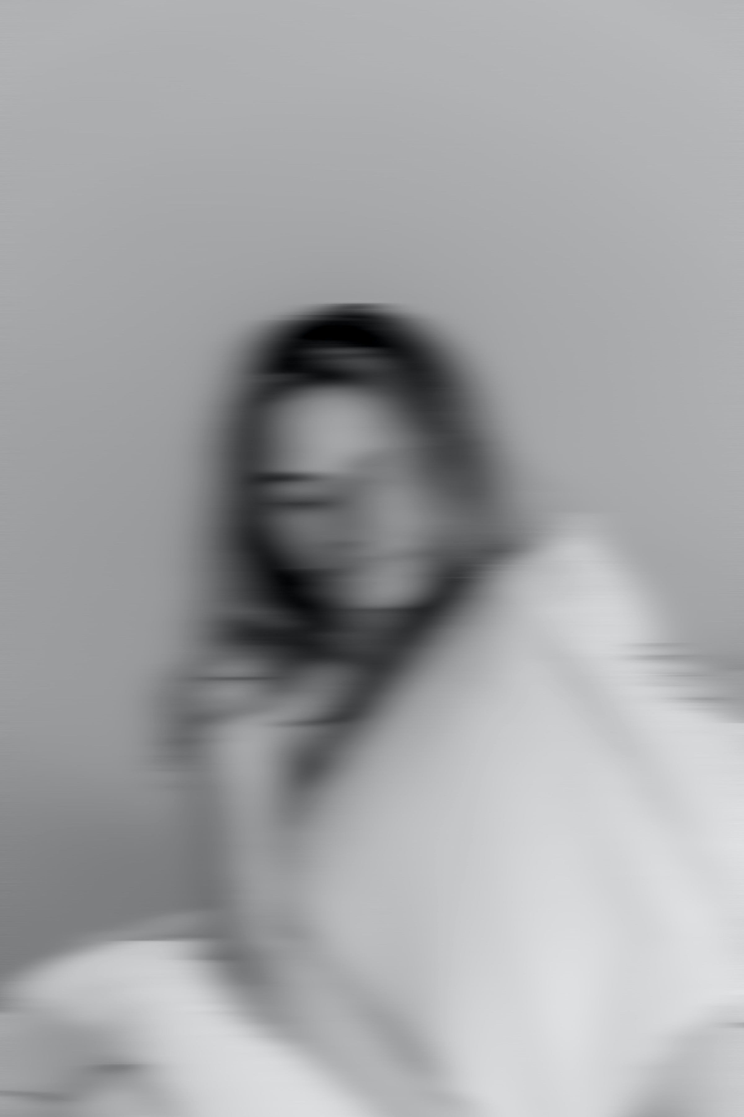 Blurred black-and-white image of a woman with dark hair, wearing a light-colored top, against a plain background.