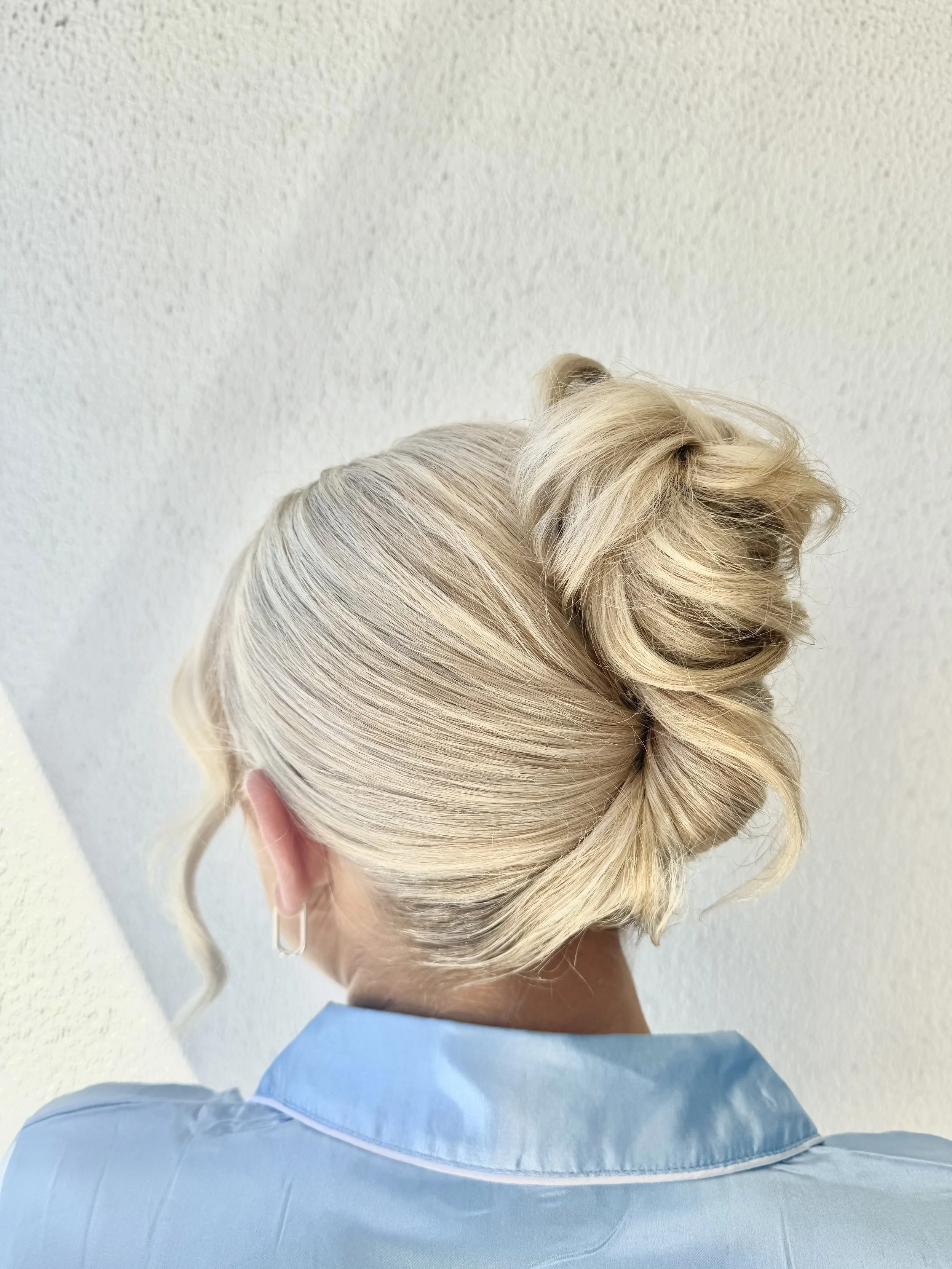 Woman with styled platinum blonde hair in an updo, wearing a light blue top and silver hoop earrings, facing away from the camera.