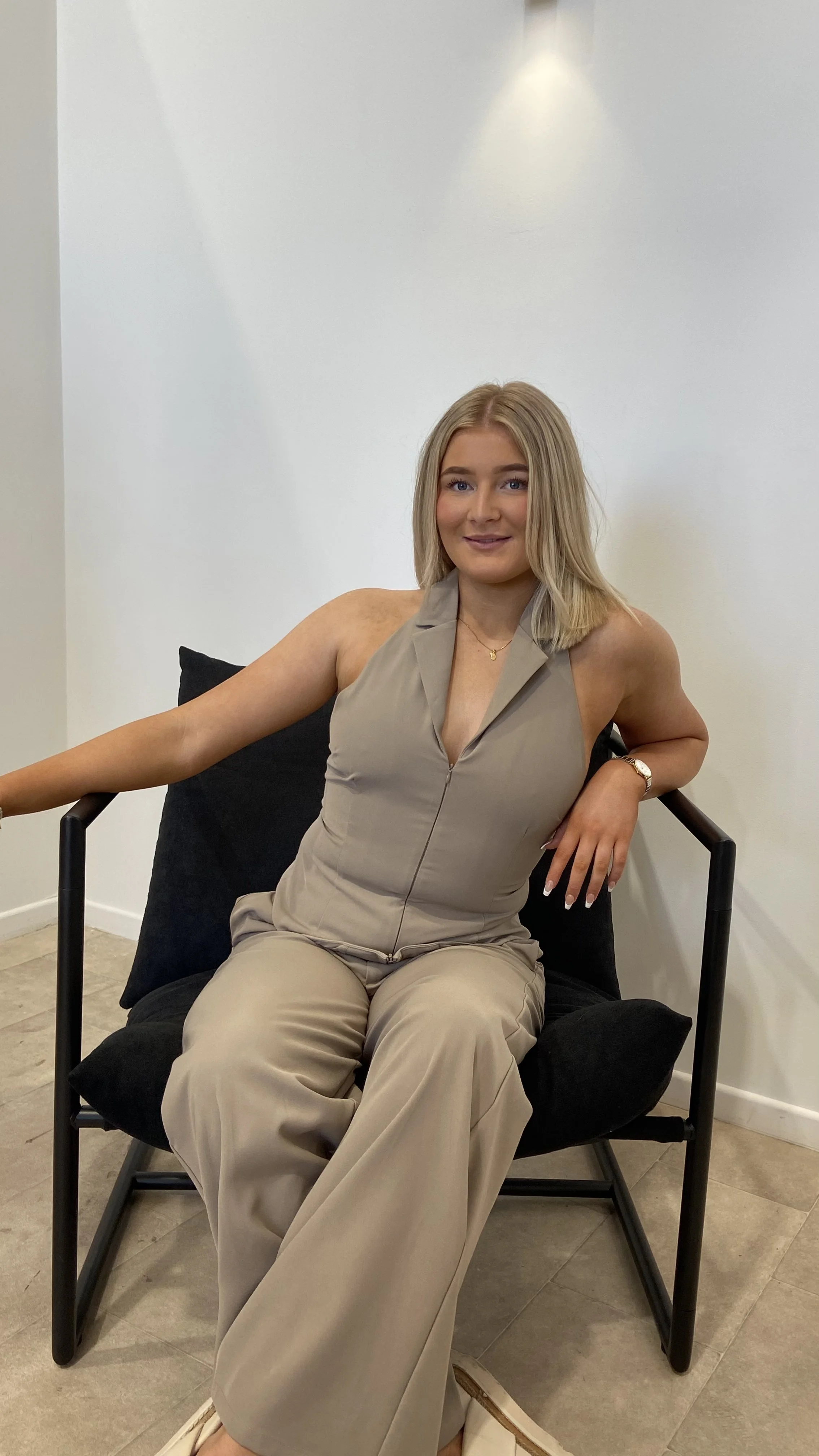 A young woman with shoulder-length blonde hair, wearing a beige sleeveless jumpsuit, sits casually on a chair with black cushions in a minimalistic room with white walls and tile flooring. She is smiling and looking at the camera.