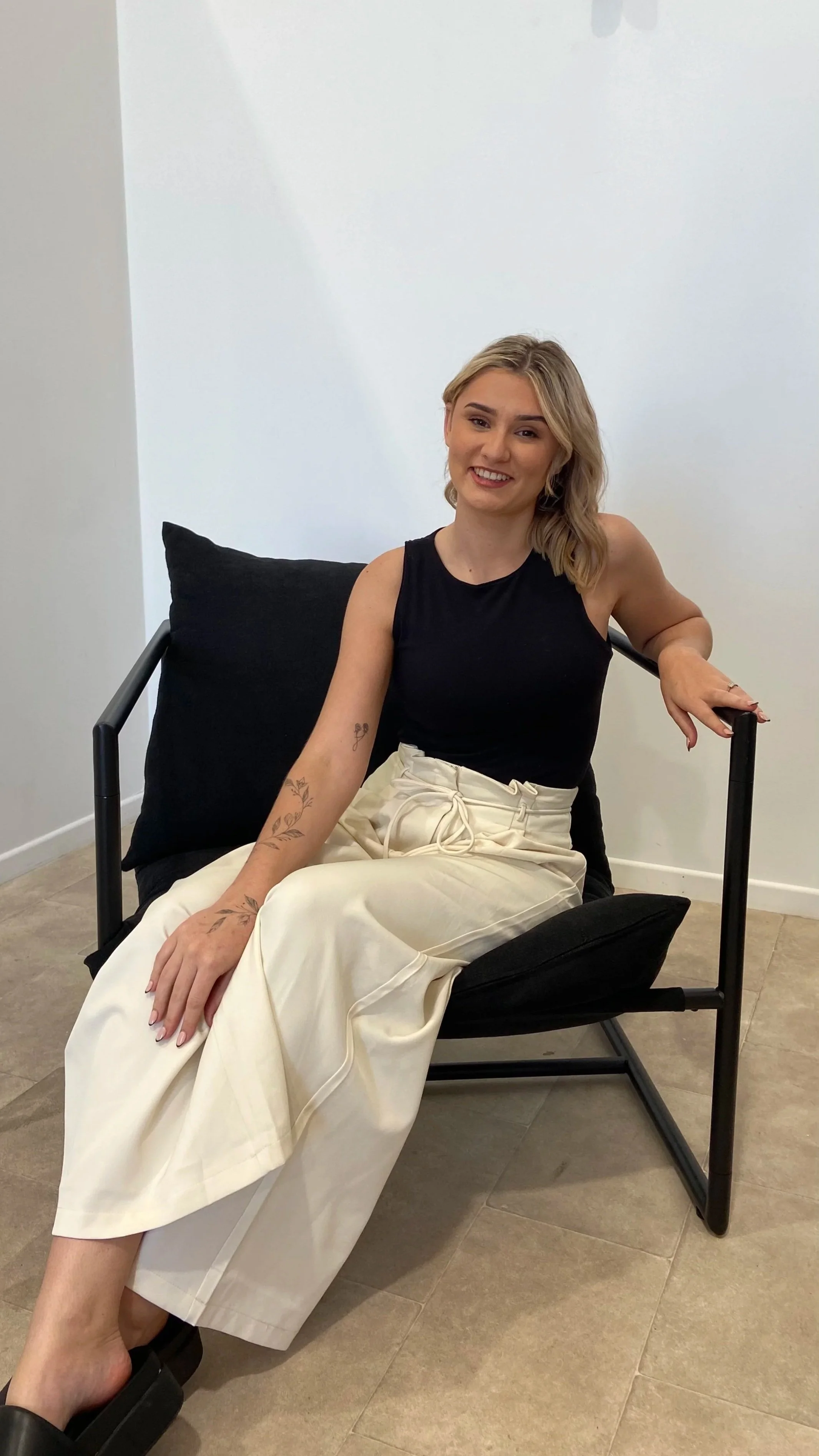 A smiling young woman with blonde hair sitting on a black chair. She is wearing a black sleeveless top and cream-colored wide-leg pants, with tattoos on her arms. She is in a minimalistic room with white walls and tiled flooring.