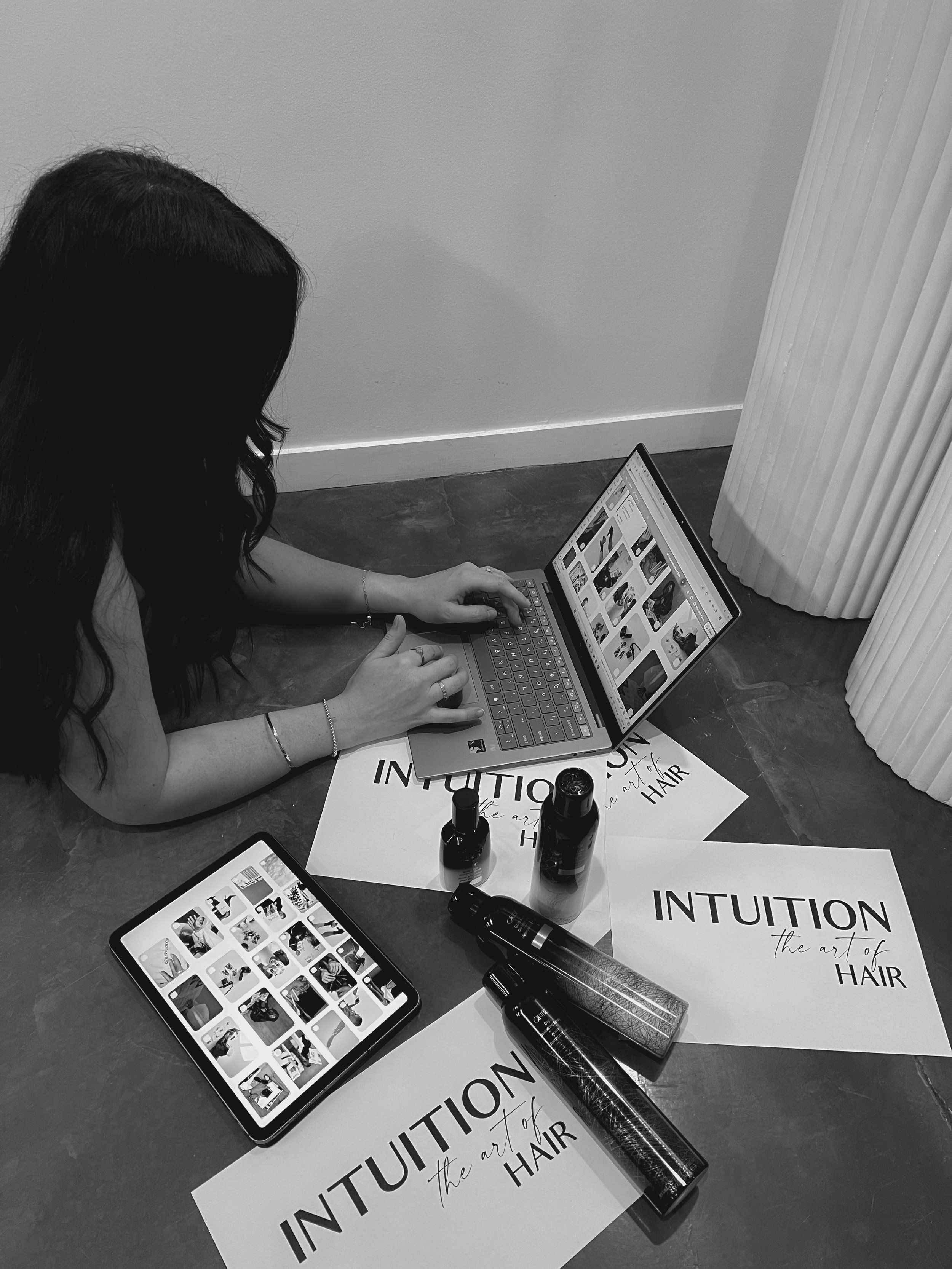 A woman with long dark hair sitting on the floor using a laptop. There are printed papers, a tablet, and bottles of hair products around her, suggesting a hair styling or hair art session.