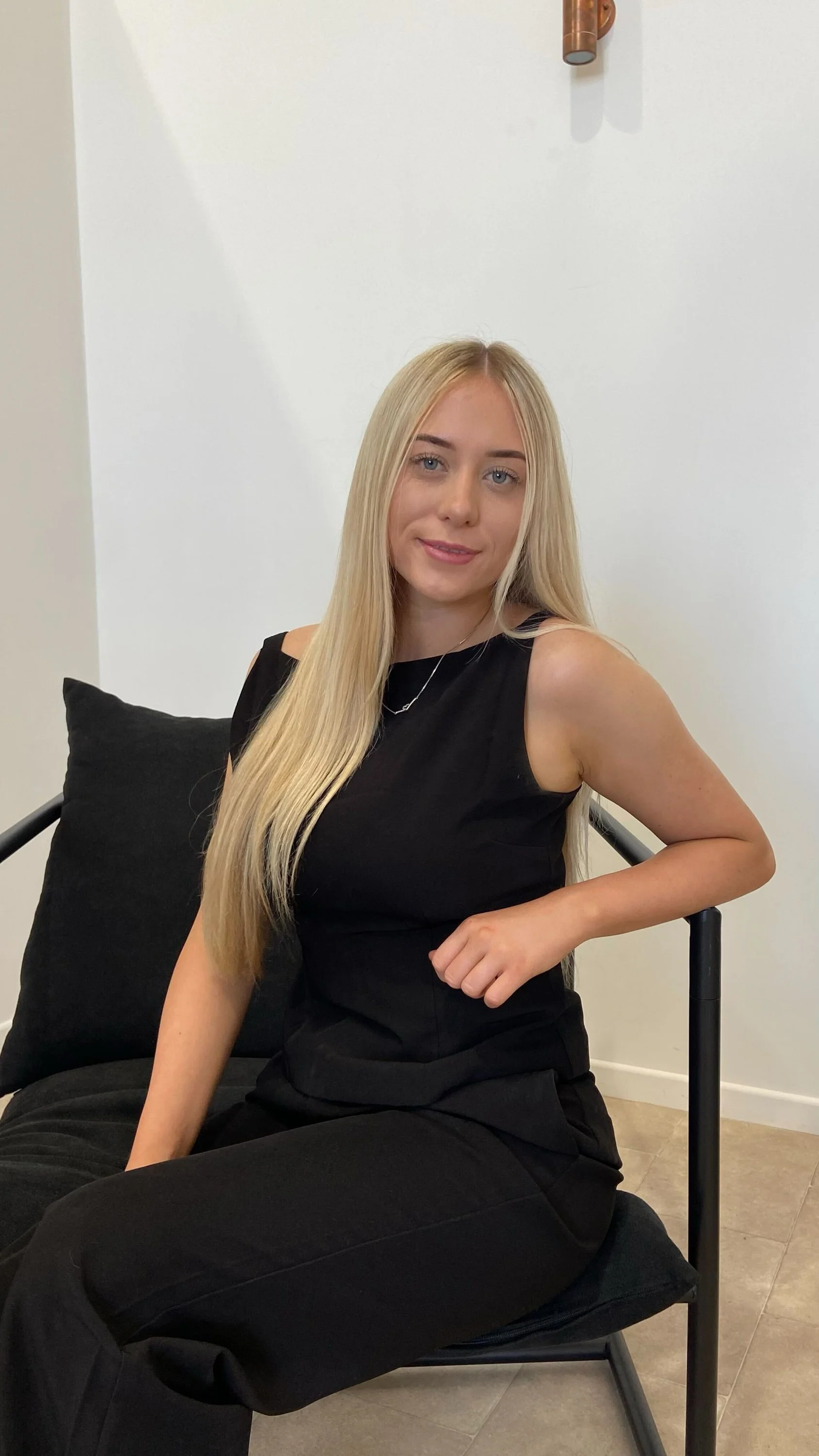 A young woman with long blonde hair, sitting on a black chair with a black pillow, wearing a black sleeveless top and pants, smiling at the camera.