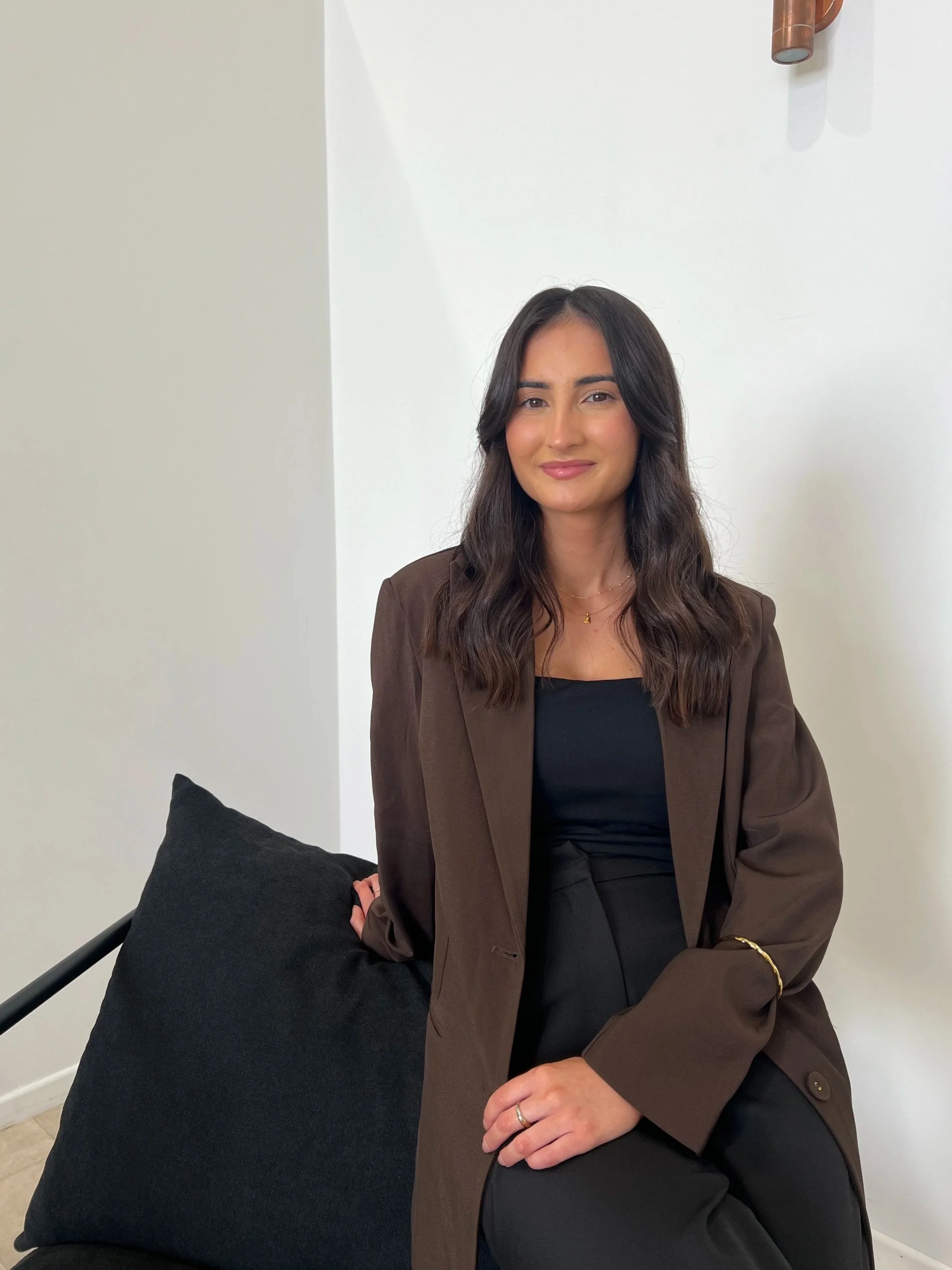 A woman with long dark hair and a gentle smile, wearing a brown blazer over a black top, sitting on a black cushion with a white wall in the background.