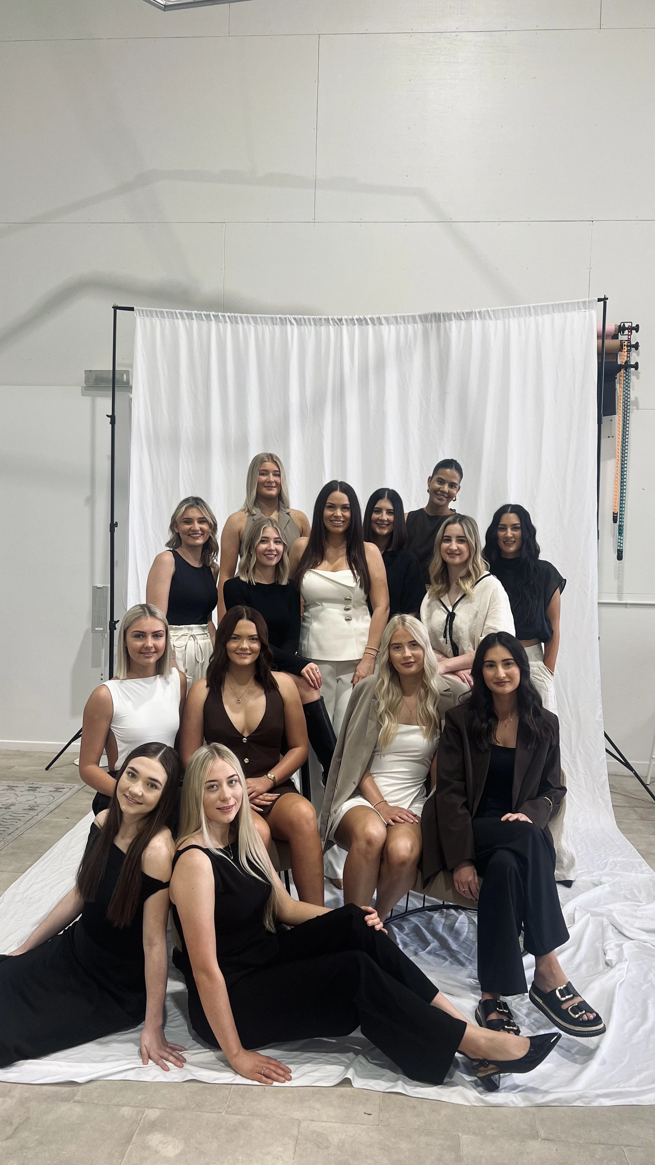 A group of 14 women posing for a photo in front of a white backdrop, some seated and some standing, dressed in fashionable black, white, beige, and brown outfits, indoors with studio lighting.
