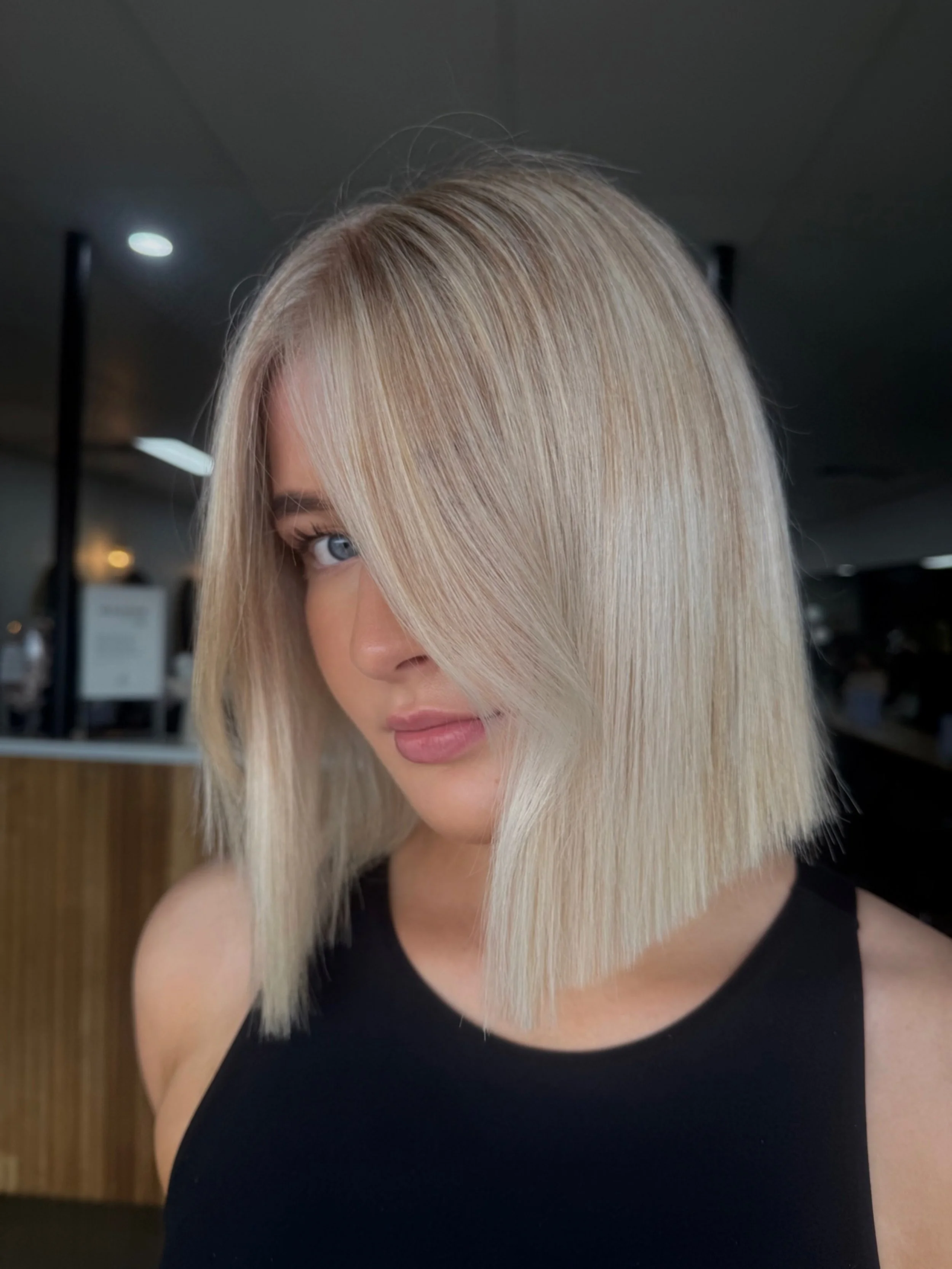 A woman with straight, shoulder-length blonde hair with purple highlights, partially covering one eye, wearing a black top, and standing indoors with blurred background.
