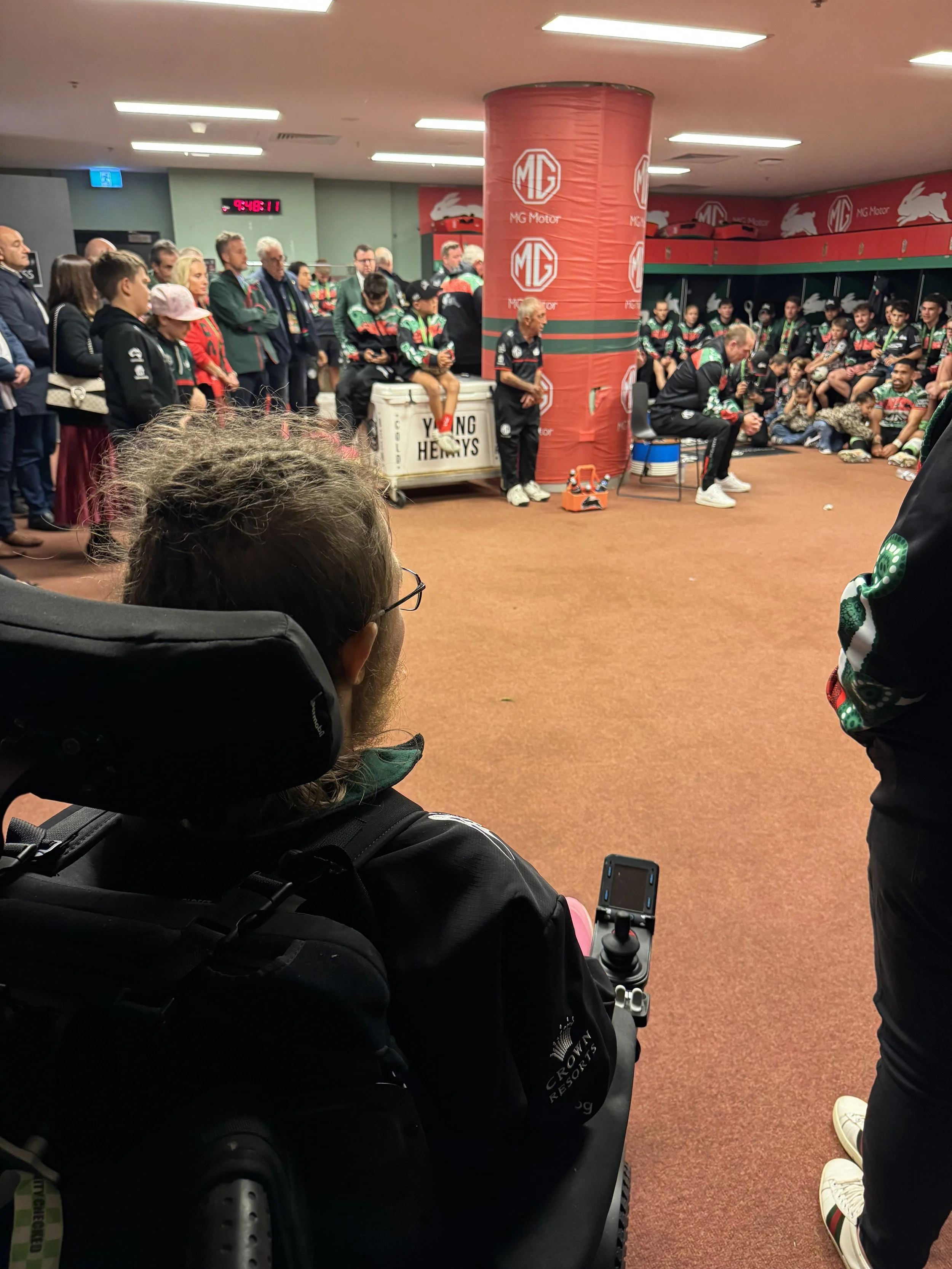 In a sports team locker room, a coach or team member speaks to a group of athletes and spectators gathered around. The room is decorated with MG Motor and Lions branding, and some individuals are seated on benches or standing.