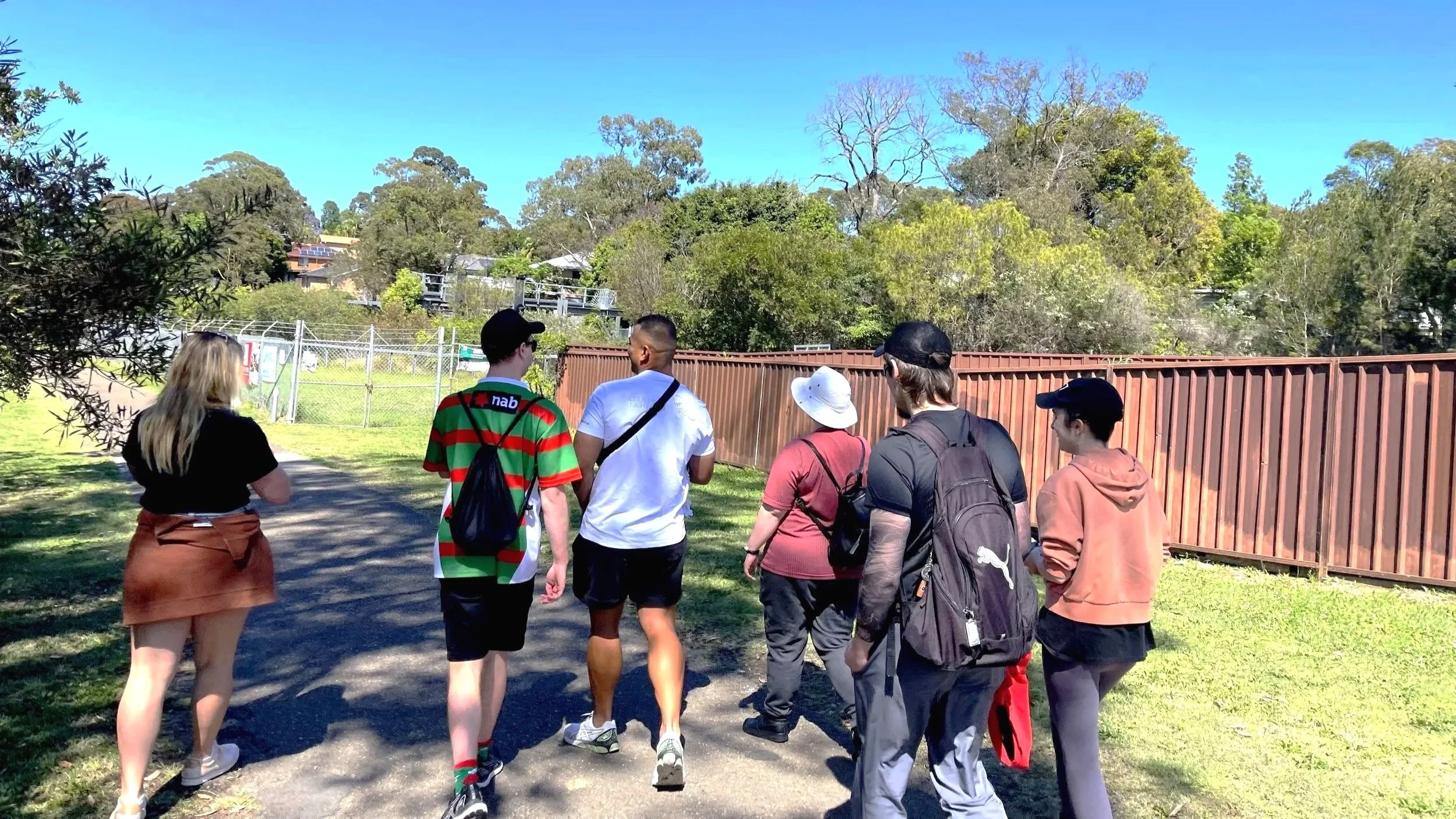 Group of seven people walking on a paved path in a park with green trees and a brown fence.