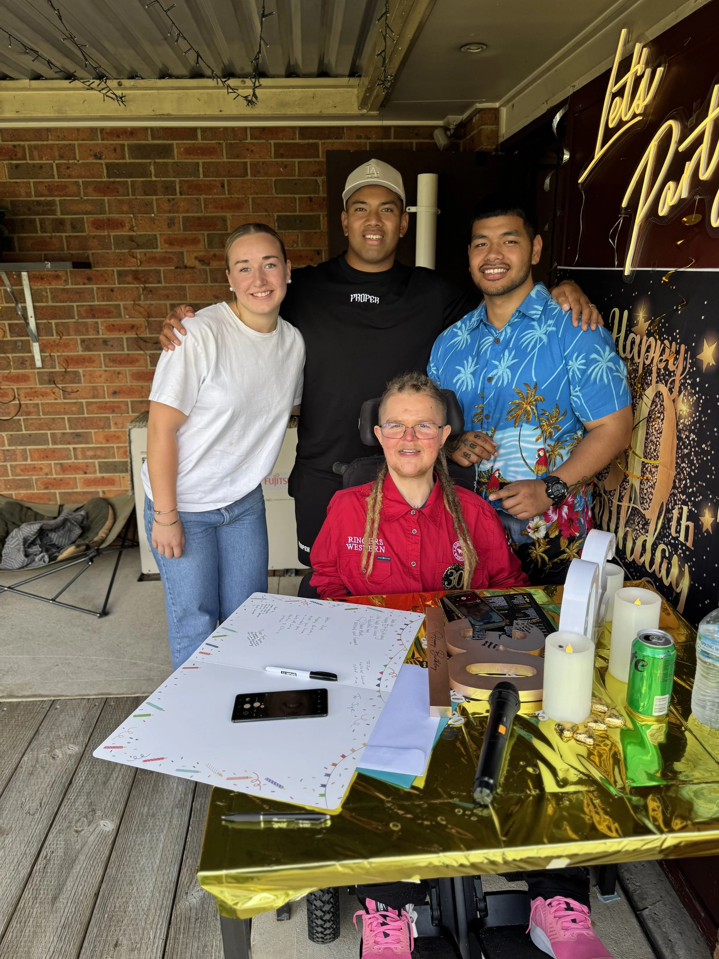 A woman in a wheelchair, wearing a red shirt, is posed with four young adults, two men and one woman, behind her, at a birthday celebration. The table in front has candles, a microphone, and birthday cards. The background includes a brick wall and a sign with gold lettering.