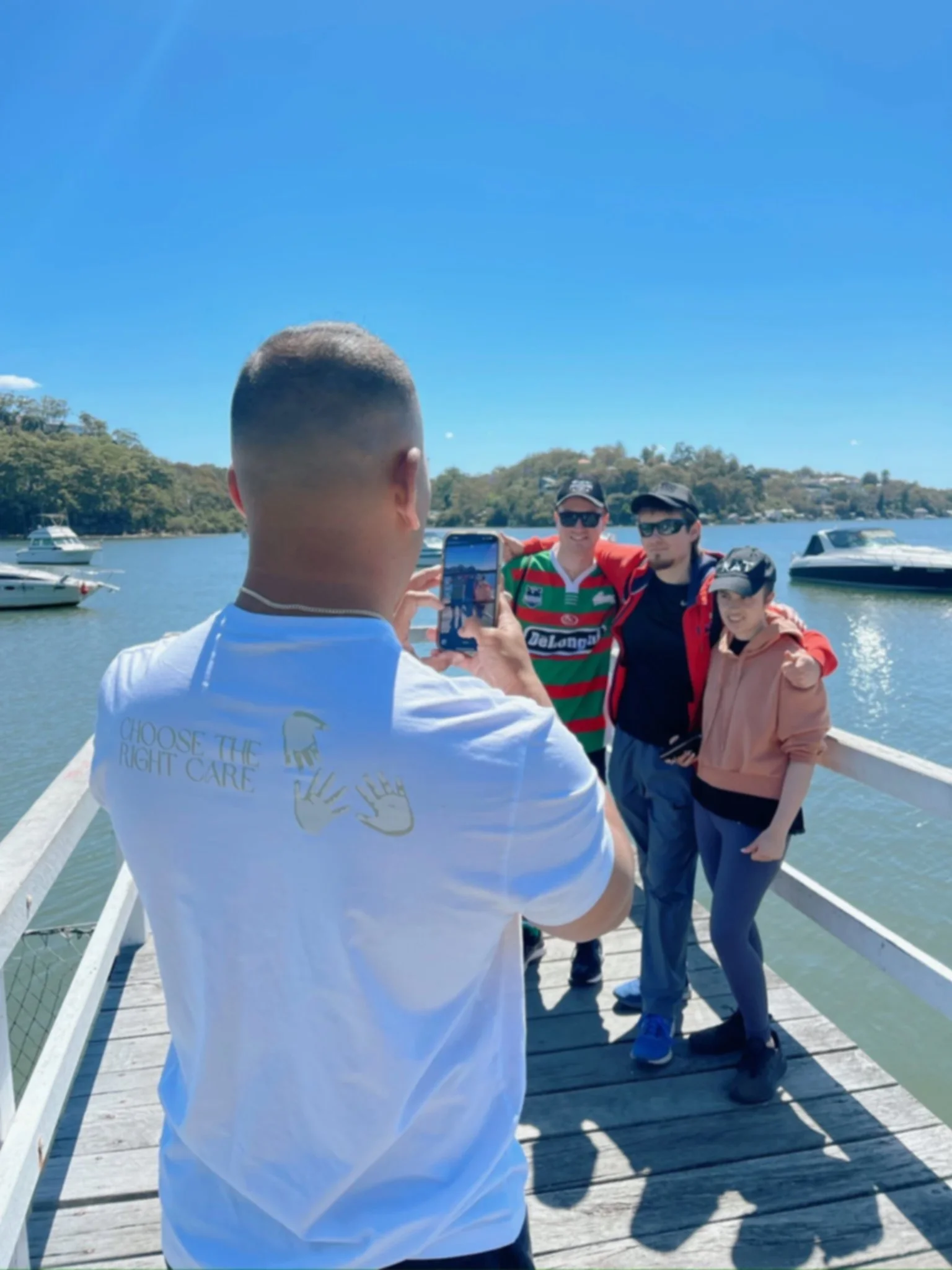 A man takes a photo of three people on a boat dock by the water with boats in the background on a sunny day.
