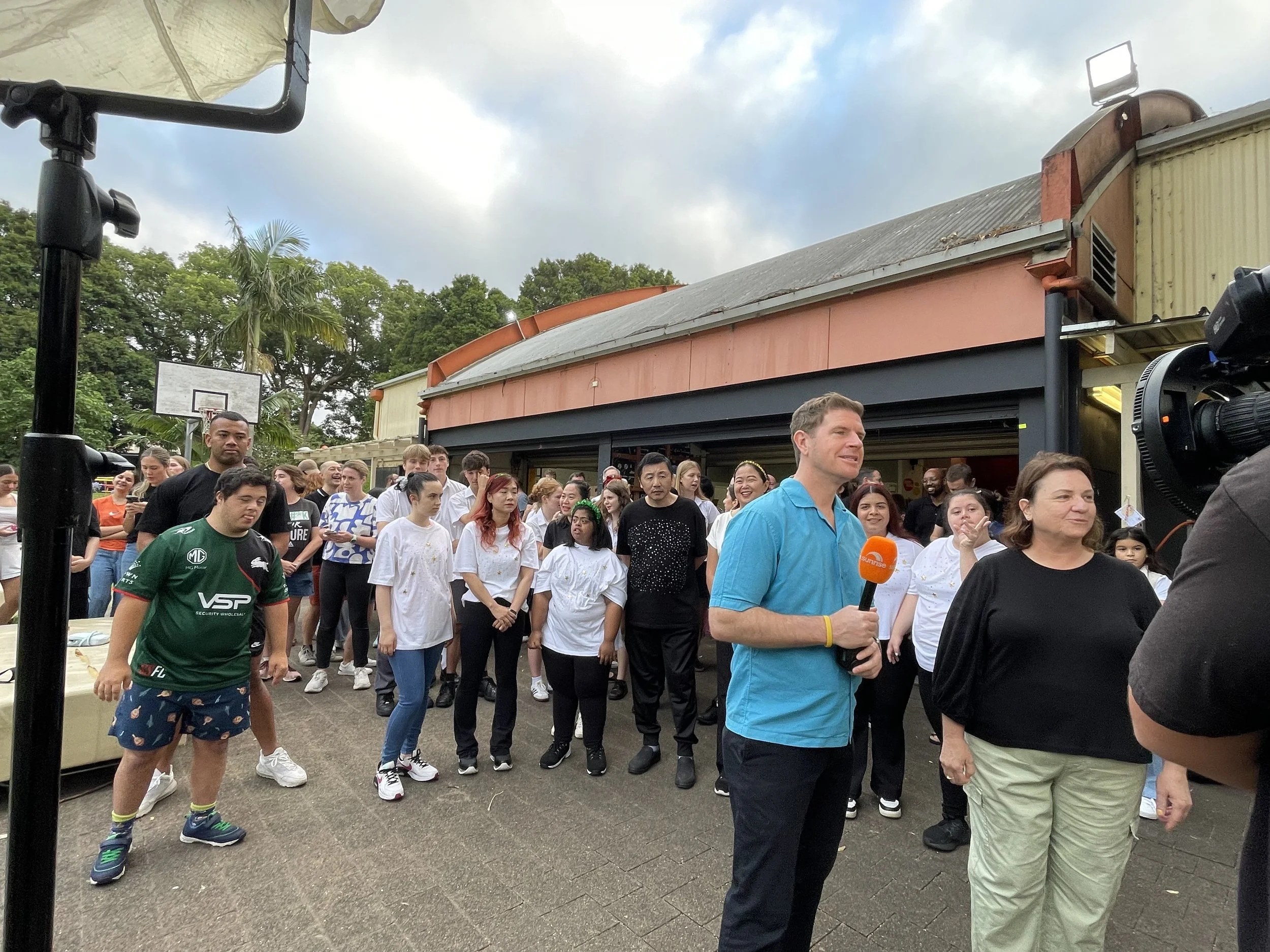 Group of people gathered outdoors for a news interview. One man in a blue shirt is holding a microphone, speaking, with a woman in black standing beside him. Several others stand behind them, some wearing white shirts. There is a basketball hoop and a colorful building in the background, with trees and cloudy sky overhead.