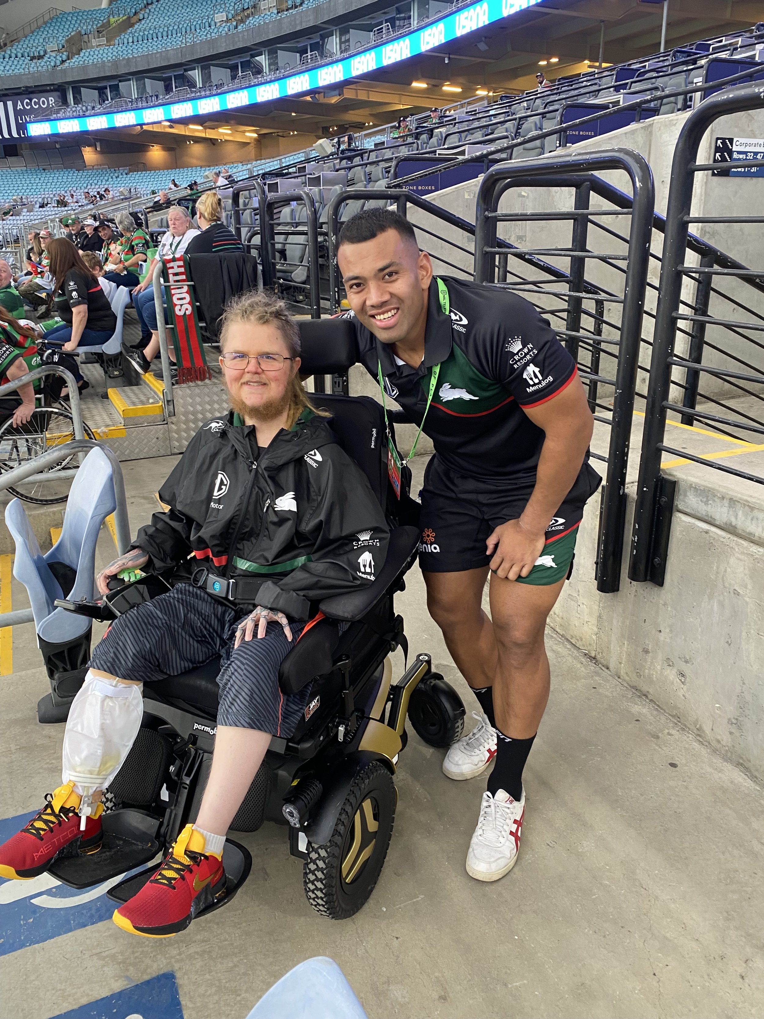A person in a wheelchair and a rugby player at a sports stadium. The person in the wheelchair is wearing a black jacket and red sports shoes, and the rugby player is dressed in a black and green uniform with white shoes, smiling at the camera.
