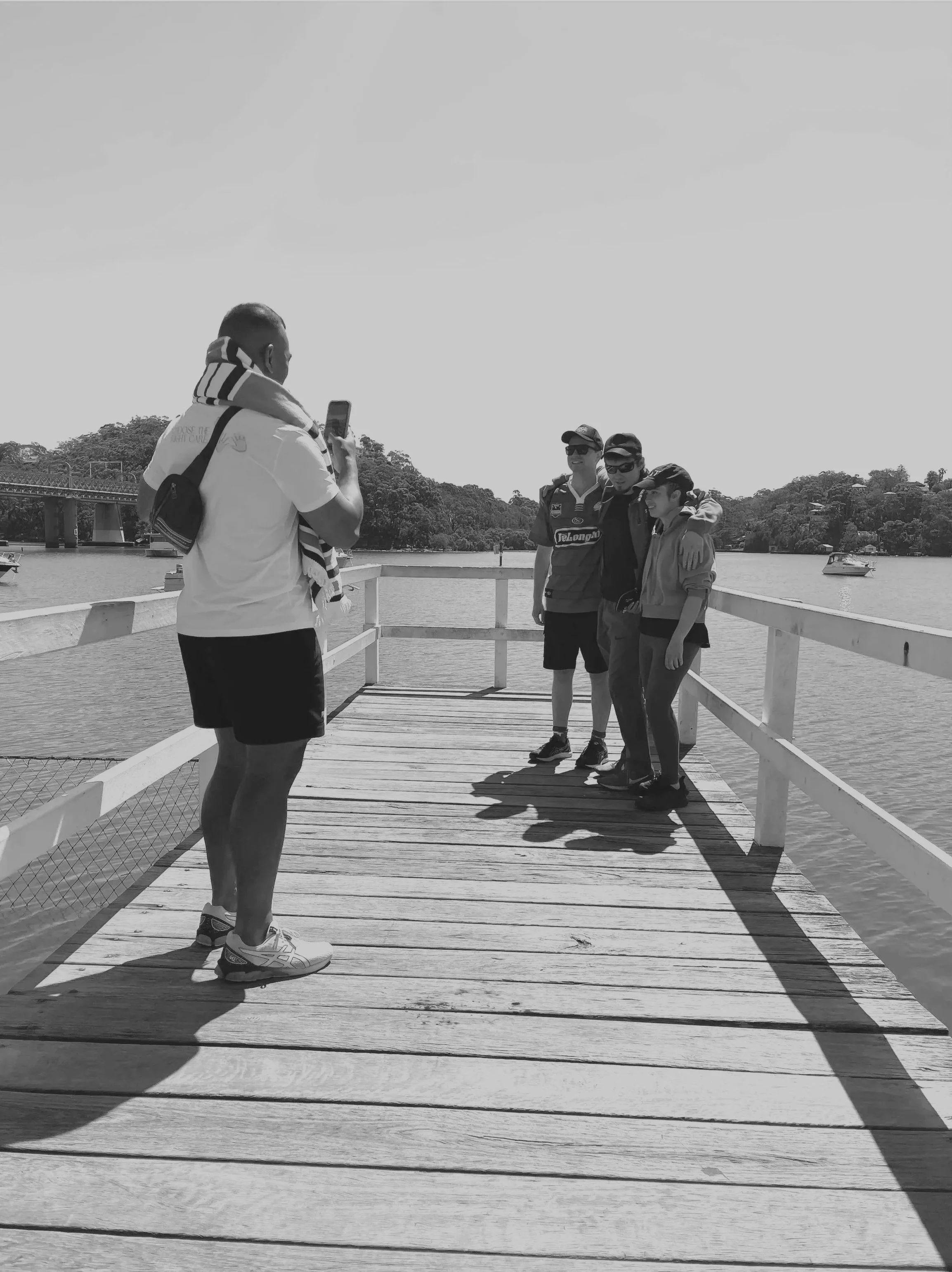 A man in a white shirt and shorts taking a photo of three people on a wooden dock by the water on a sunny day.