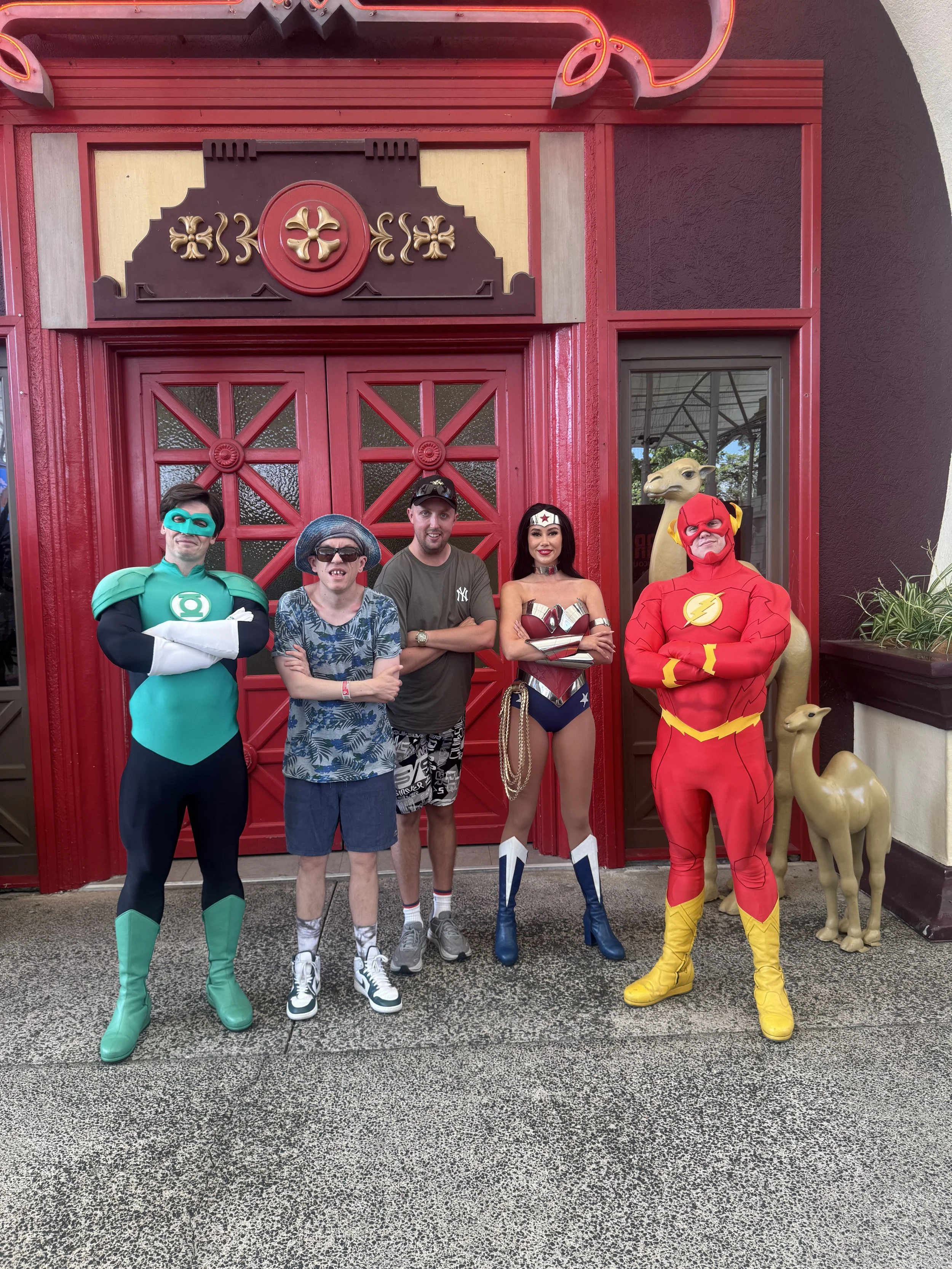 Three people dressed in casual clothing and two people in superhero costumes posing in front of a red, decorative building entrance with camel statues on each side.